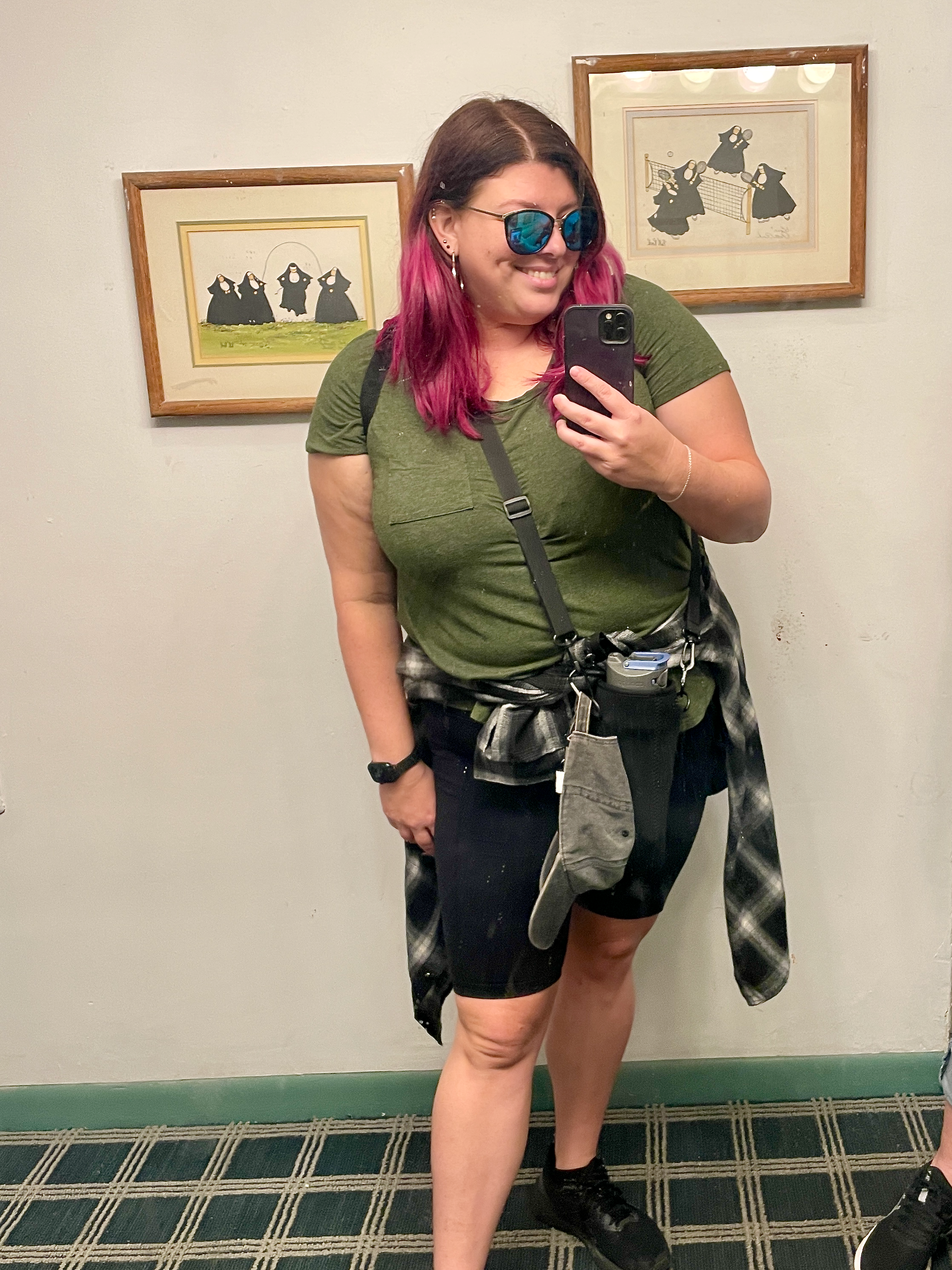 Young woman with pink and brown hair wearing sunglasses takes a mirror selfie, standing in front of framed pictures of penguins playing volleyball on a white wall.