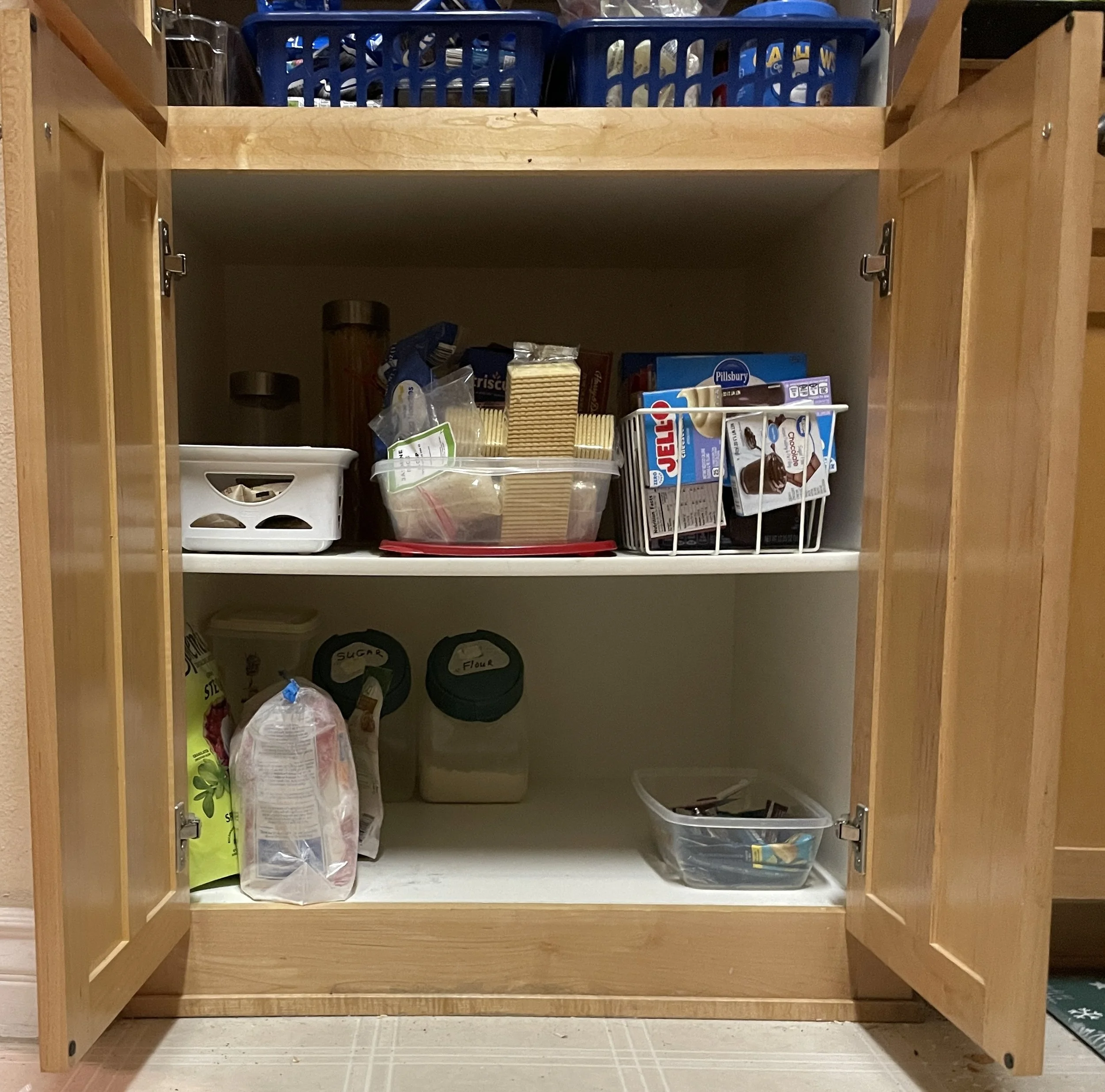 Open wooden kitchen cabinet with baking supplies, cereal boxes, and snack bags inside.