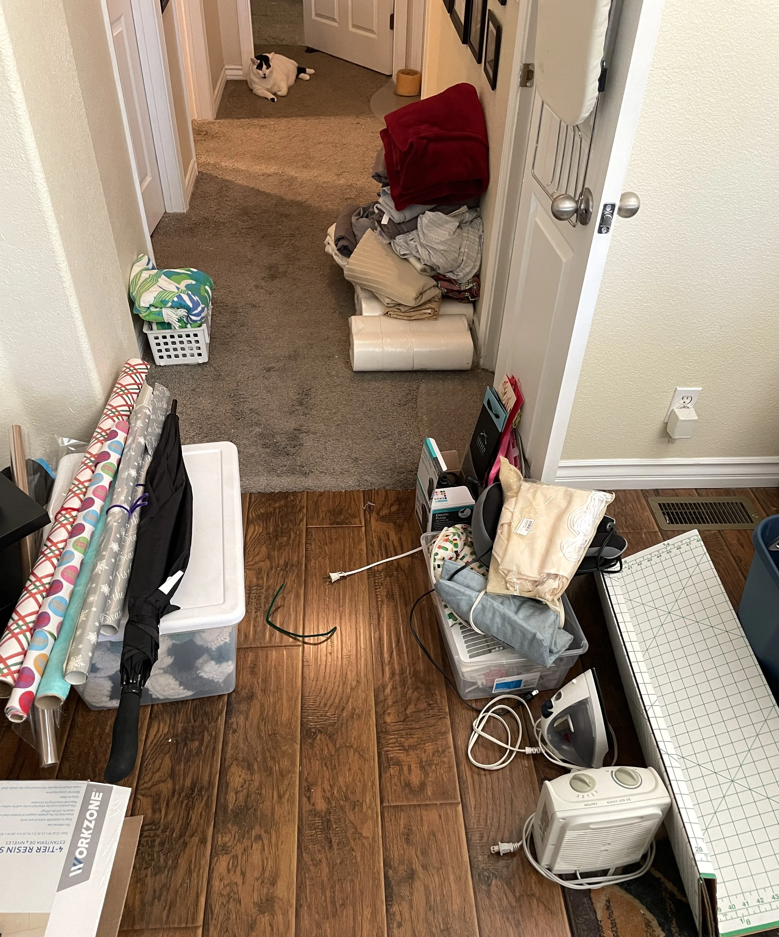 A cluttered indoor space with a small white heater, various rolls of wrapping paper, a basket with clothes, a wrapped package, and cords on a wooden floor. In the background, a hallway with a black and white cat lying on the carpet near a doorway.