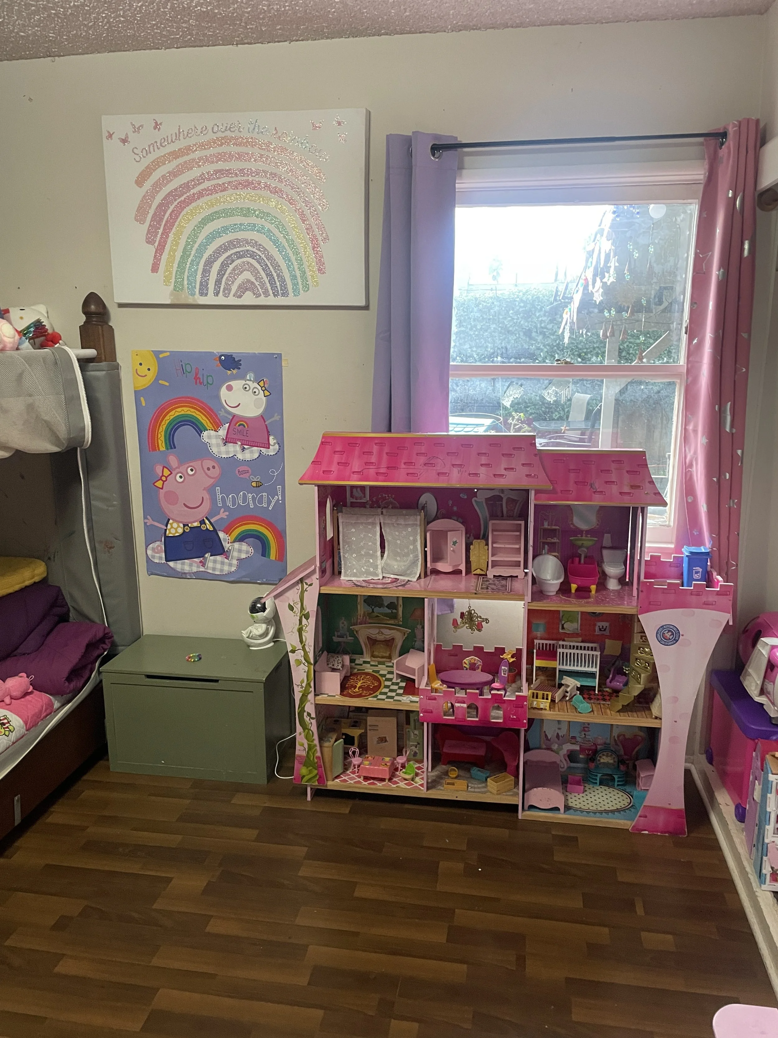 A children's playroom with a pink dollhouse, a green nightstand, and Peppa Pig and rainbow-themed posters on the wall. There are purple and pink curtains by a window, and books on a shelf on the right.
