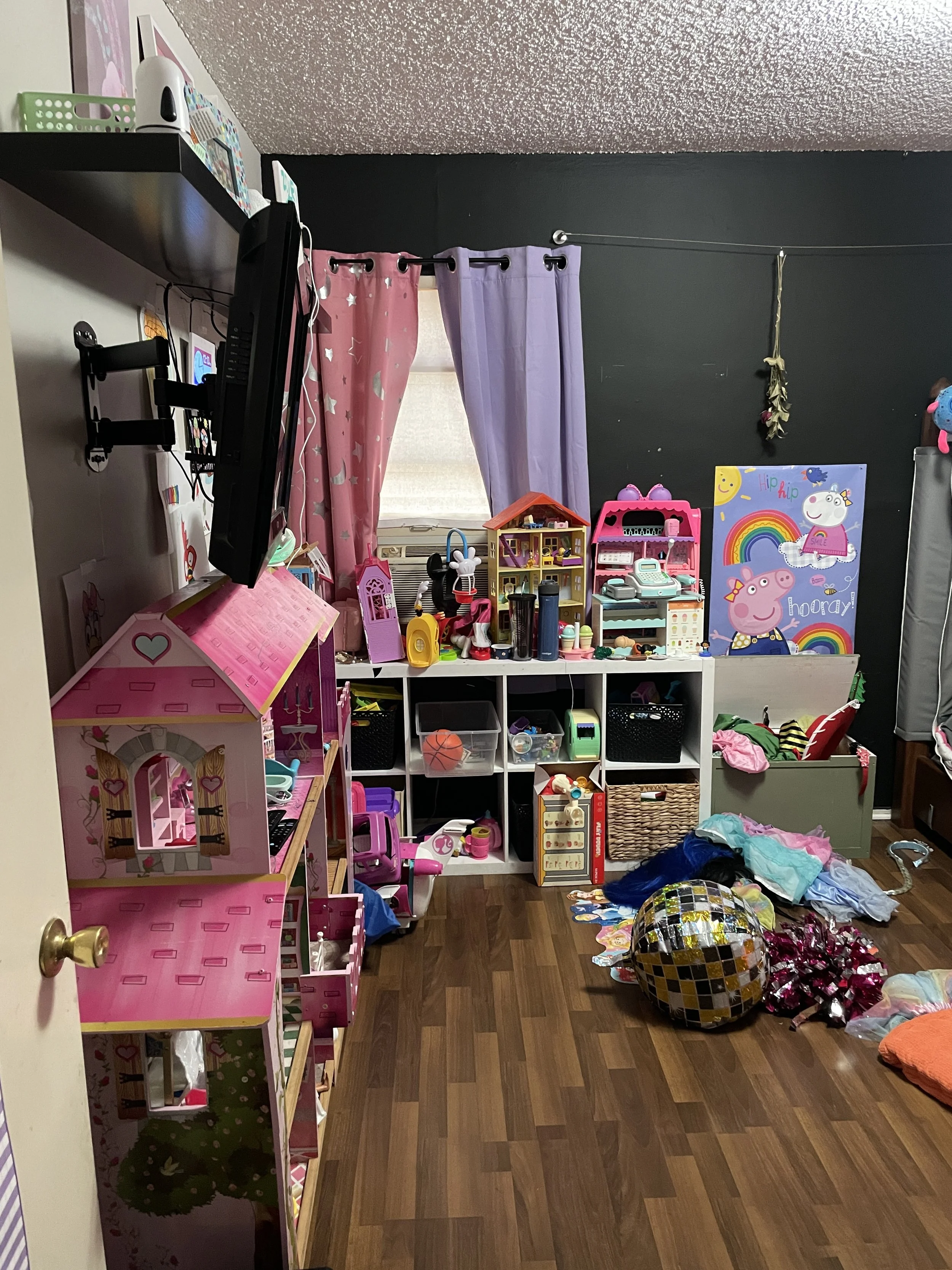 A messy children's playroom with toys, dolls, and stuffed animals scattered on the floor. Pink and purple curtains hang over a window. Shelves with various toys and a large Peppa Pig poster are visible in the background.