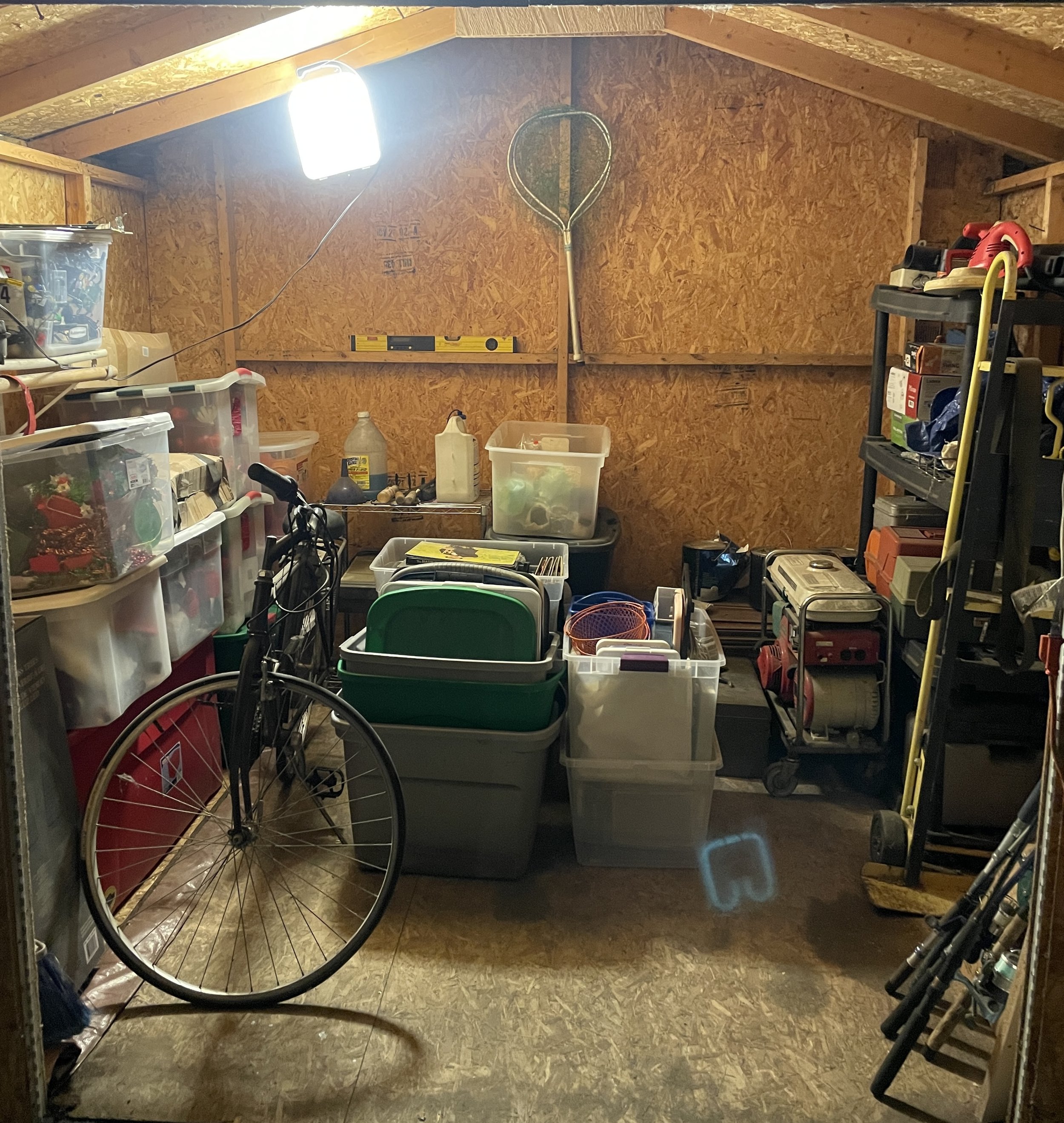 Inside a cluttered garage with various storage containers, tools, and equipment, including a wheelchair and a tennis racket hanging on the wall.