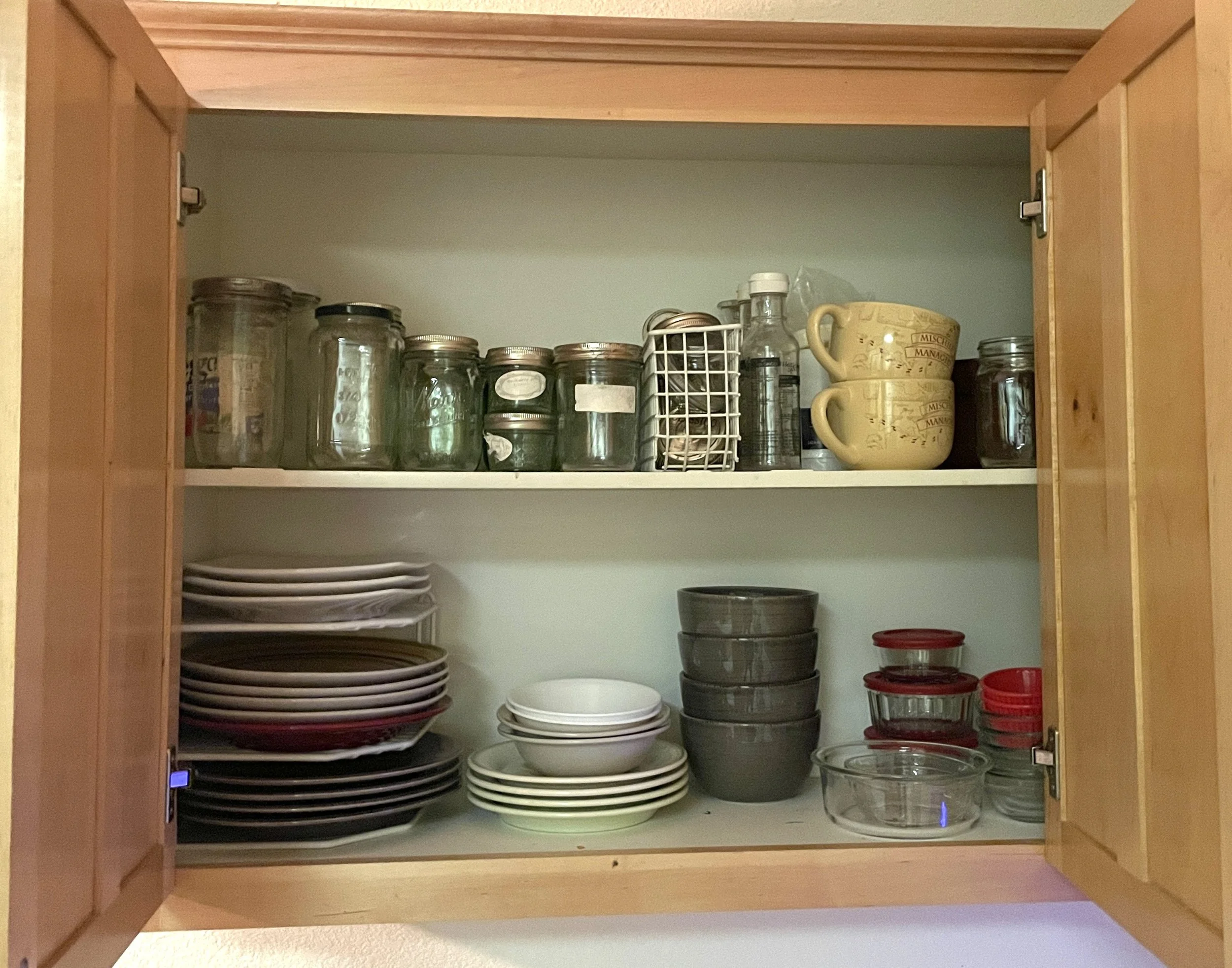 An open kitchen cabinet with two shelves containing various glass and ceramic dishes, cups, bowls, and jars.