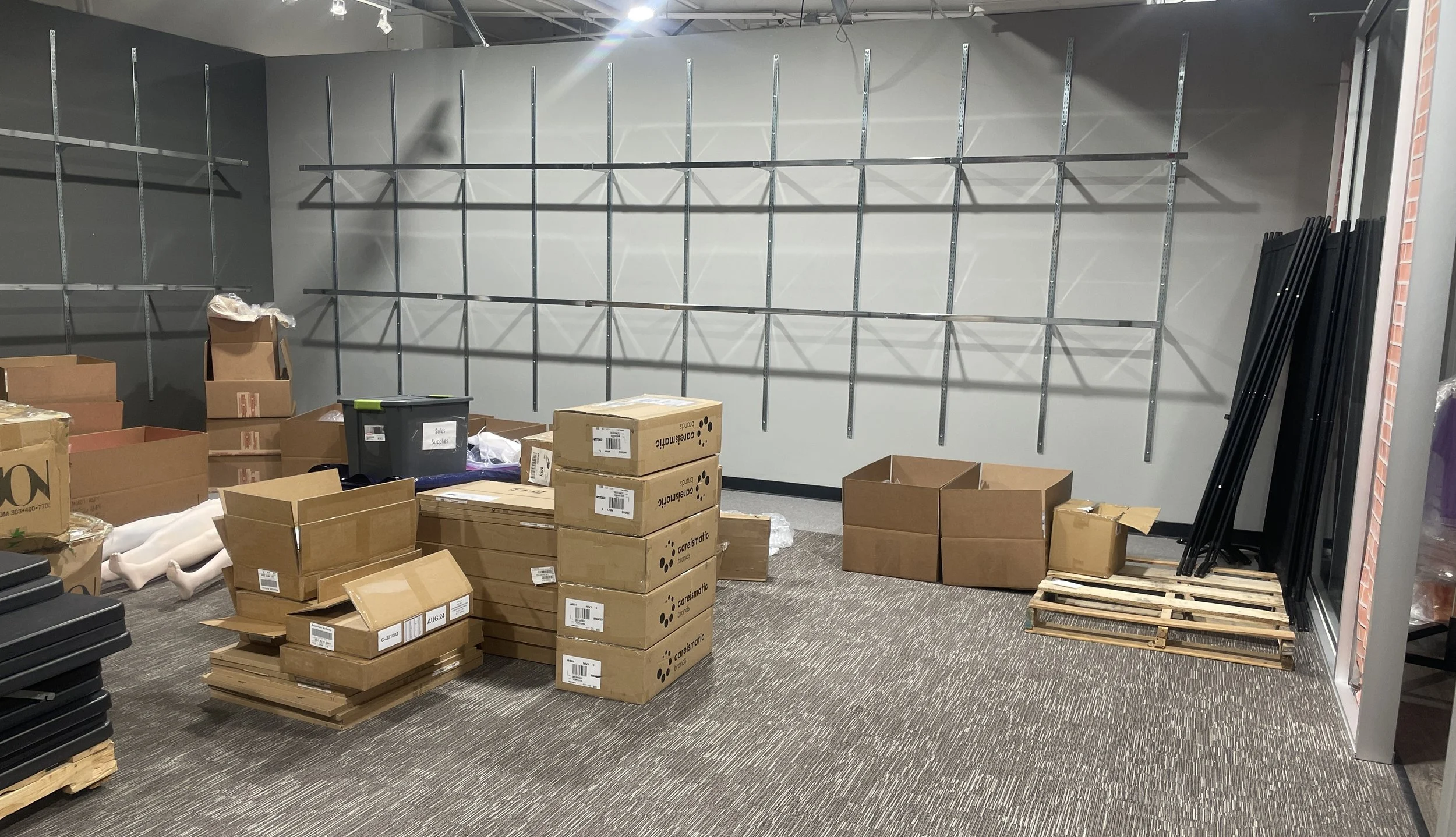 Empty retail or storage area with metal wall brackets on gray wall, boxes and pallets on carpeted floor, leaning black metal frames on the right.