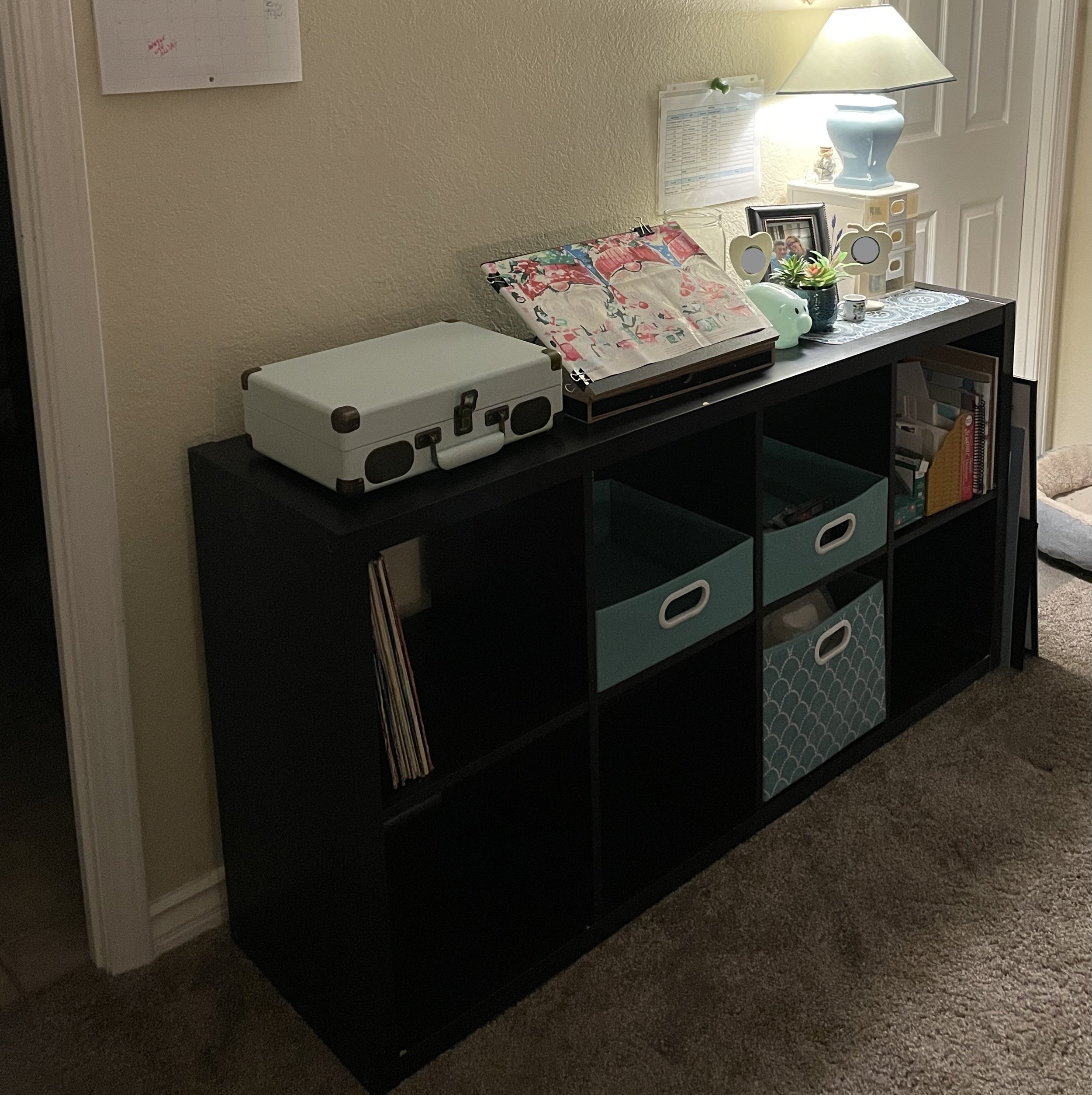A black shelving unit against a beige wall with various items on top, including a white box, photo frames, a small potted succulent, a blue piggy bank, and a table lamp. There's a colorful fabric or paper on the shelf, and binders or books below. A whiteboard is mounted on the wall above the shelf.