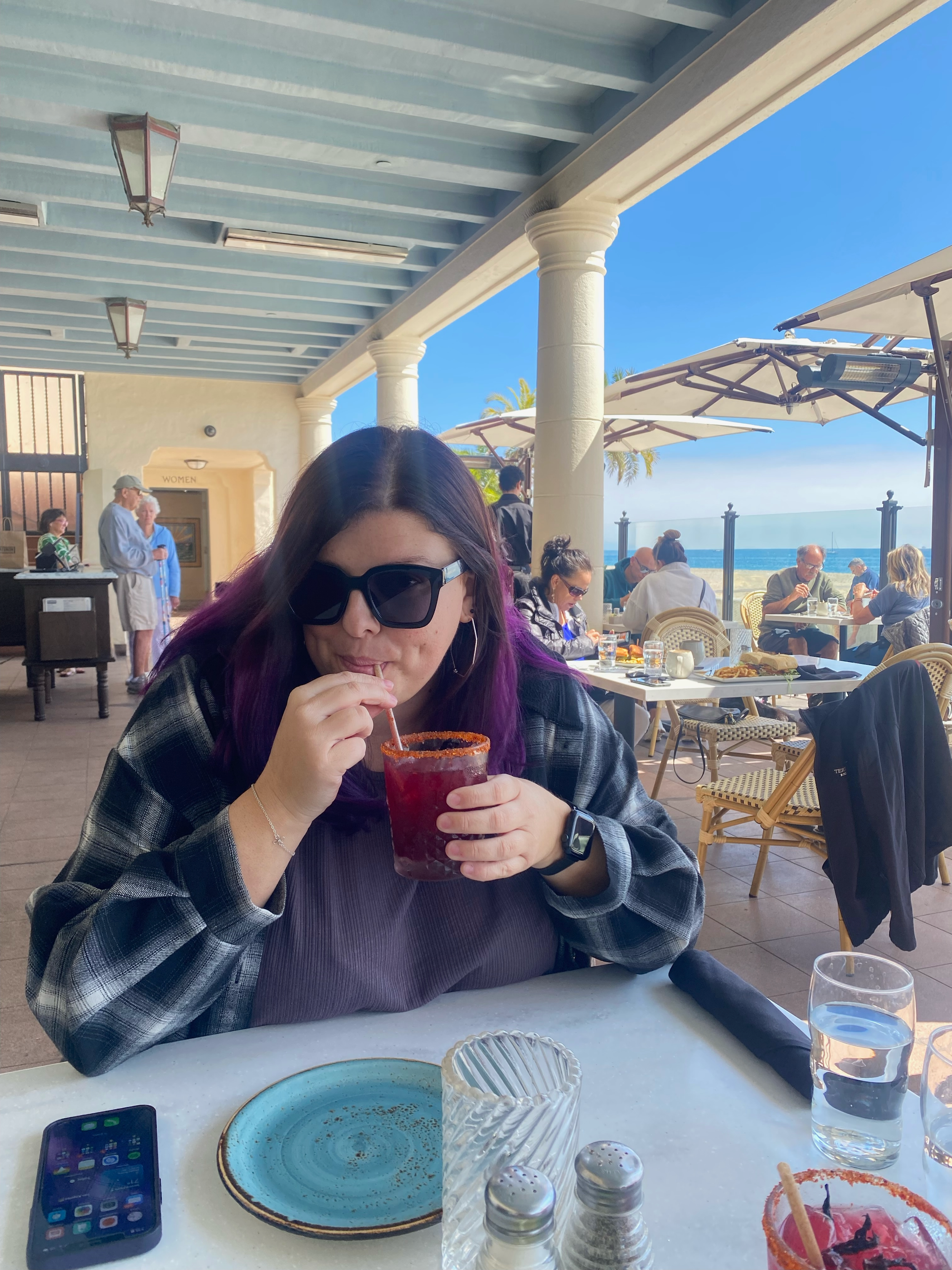 A woman with purple hair, large black sunglasses, and a black and gray checkered shirt drinking a red beverage through a straw at an outdoor restaurant with ocean view.