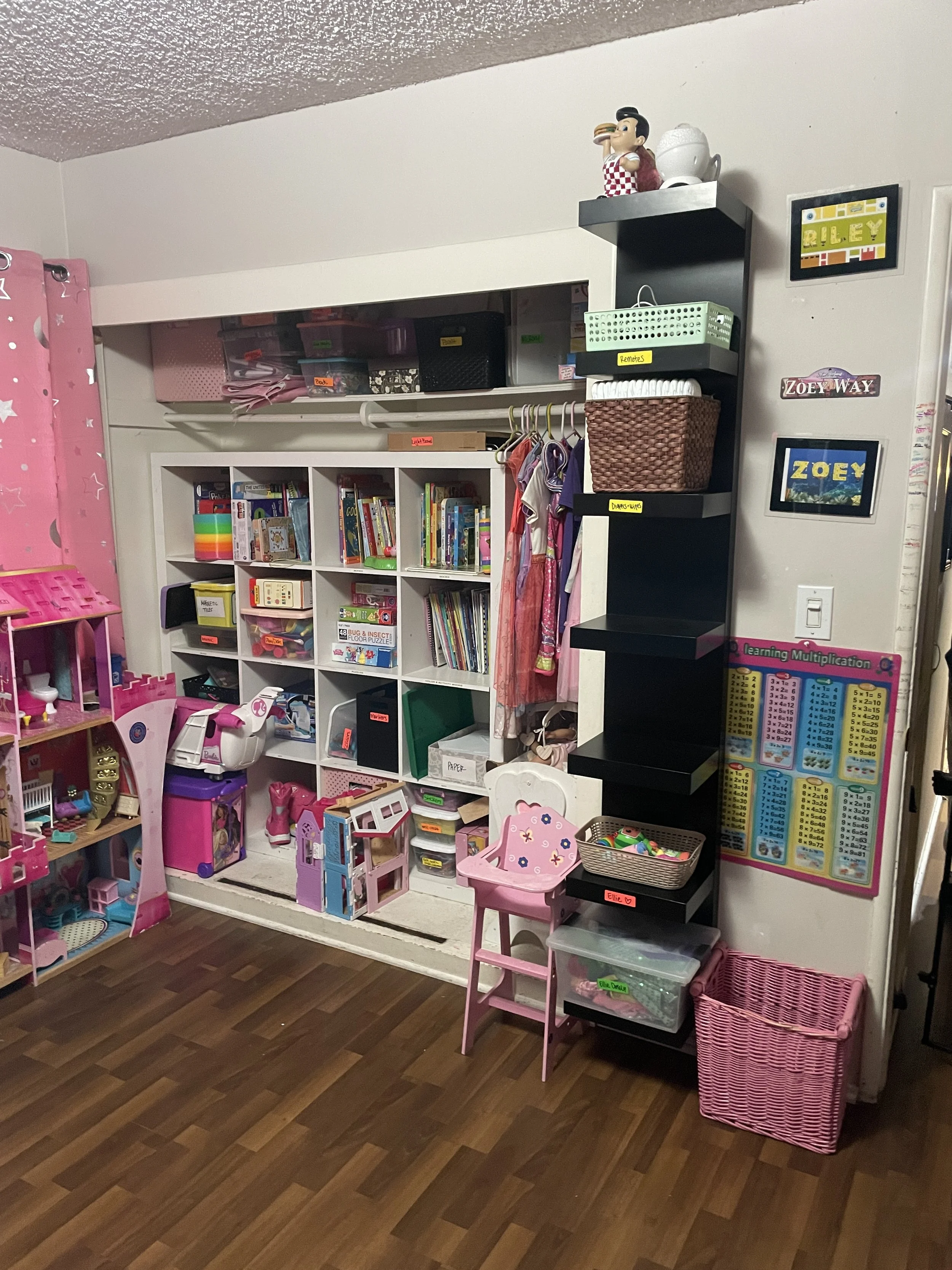 A child's playroom with bookshelves, toys, and storage bins, featuring a pink chair, a pink basket, and shelves labeled for organization.