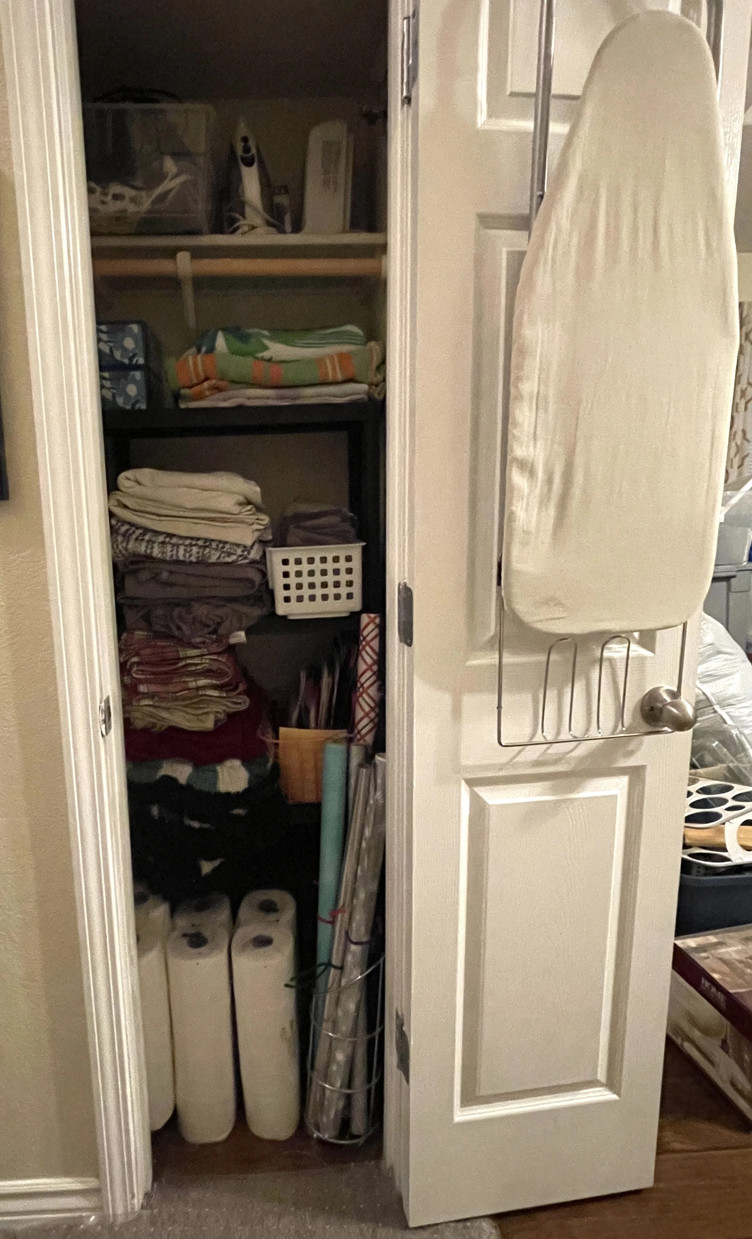 Open closet door revealing shelves with folded towels, a laundry basket, and various household items. The door has a white ironing board attached.
