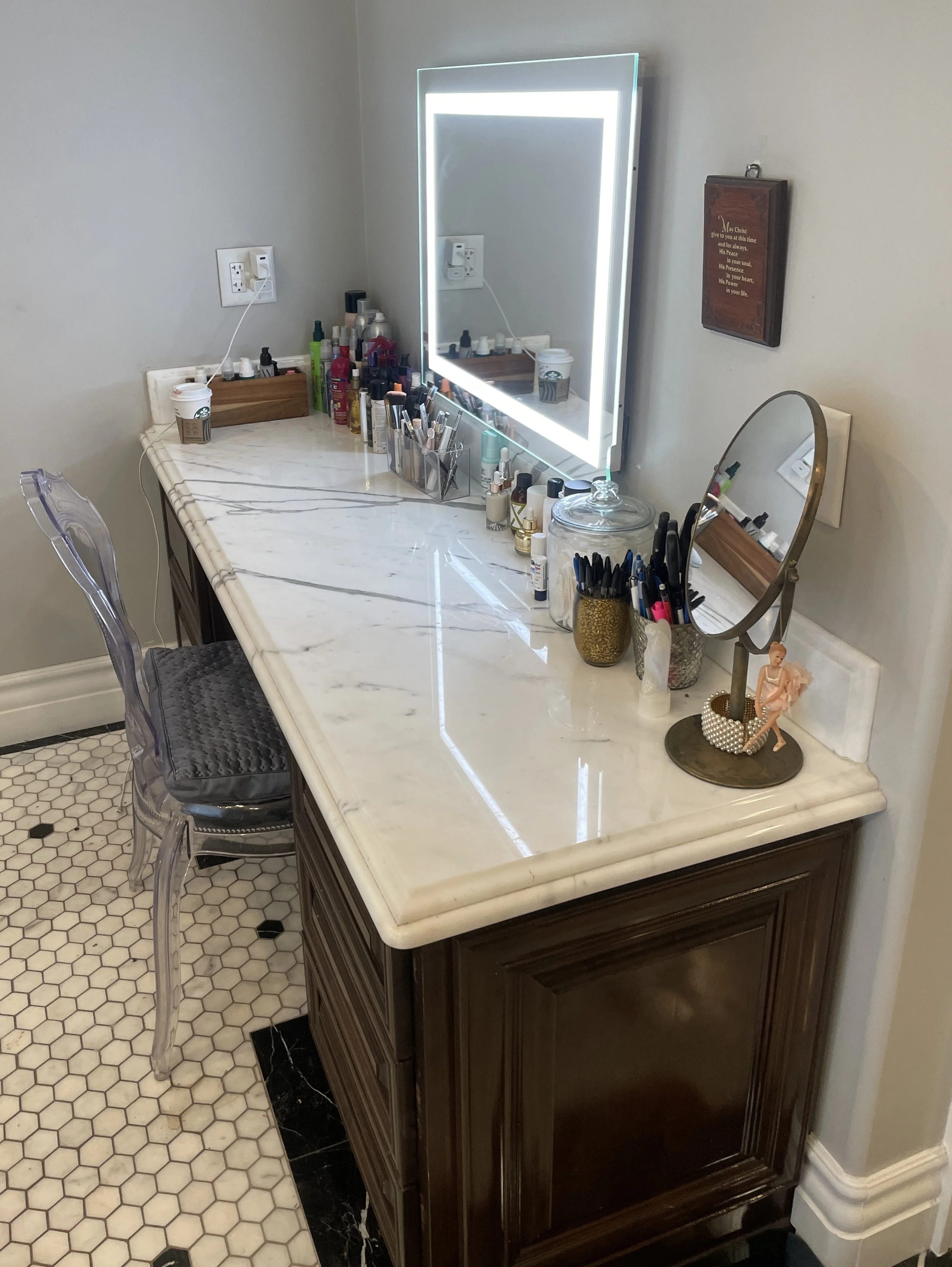 A makeup vanity with a large illuminated mirror, holding various makeup products, brushes, and a globe-shaped container. A small sculpture of a ballerina is next to a small vase, and a wooden wall plaque with a quote hangs on the wall.