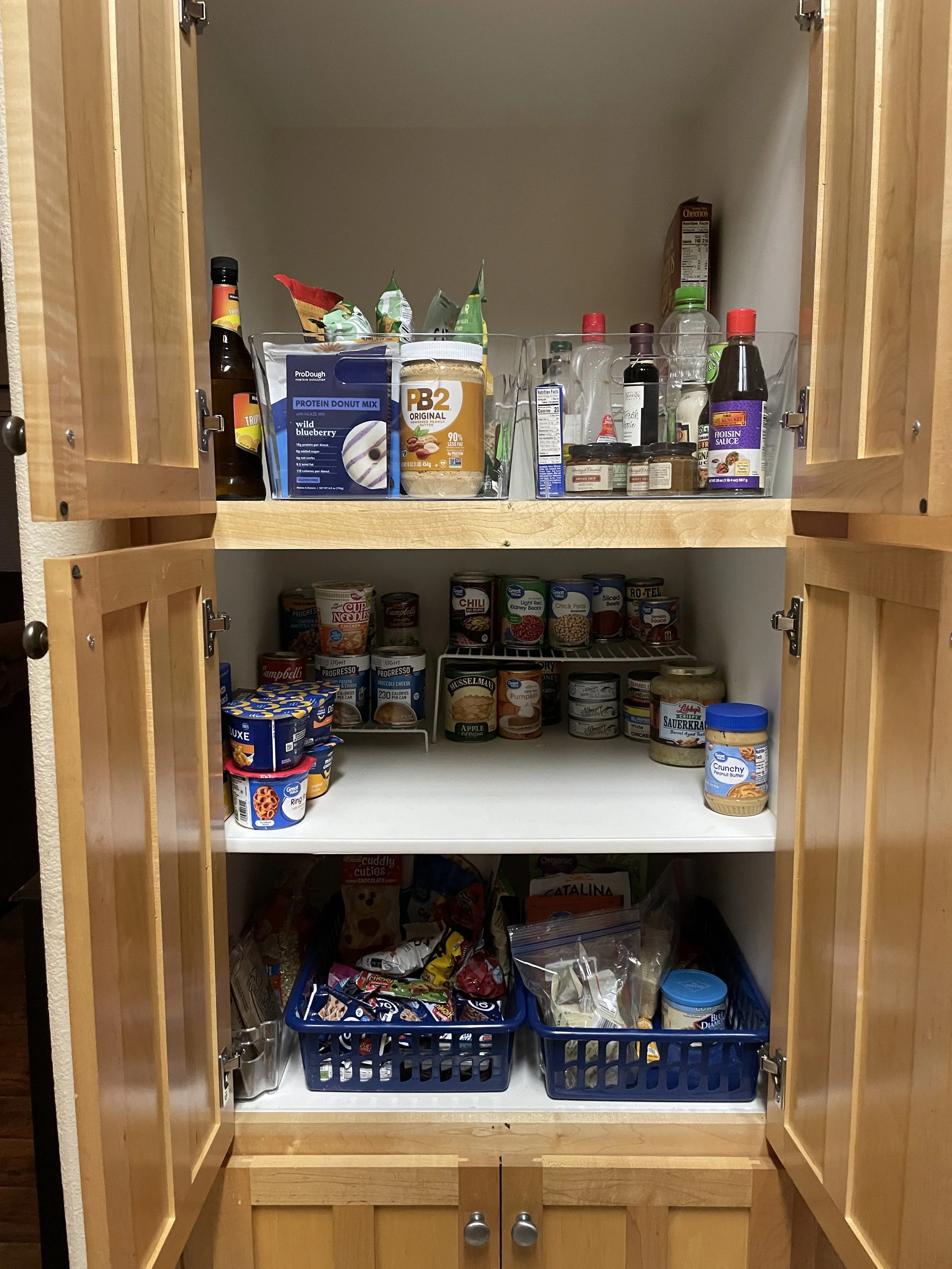 Kitchen pantry with three shelves filled with various canned goods, snacks, and beverages. The top shelf contains bottles and boxes, the middle shelf has canned foods and yogurt, and the bottom shelf holds snack bags and other packaged items.