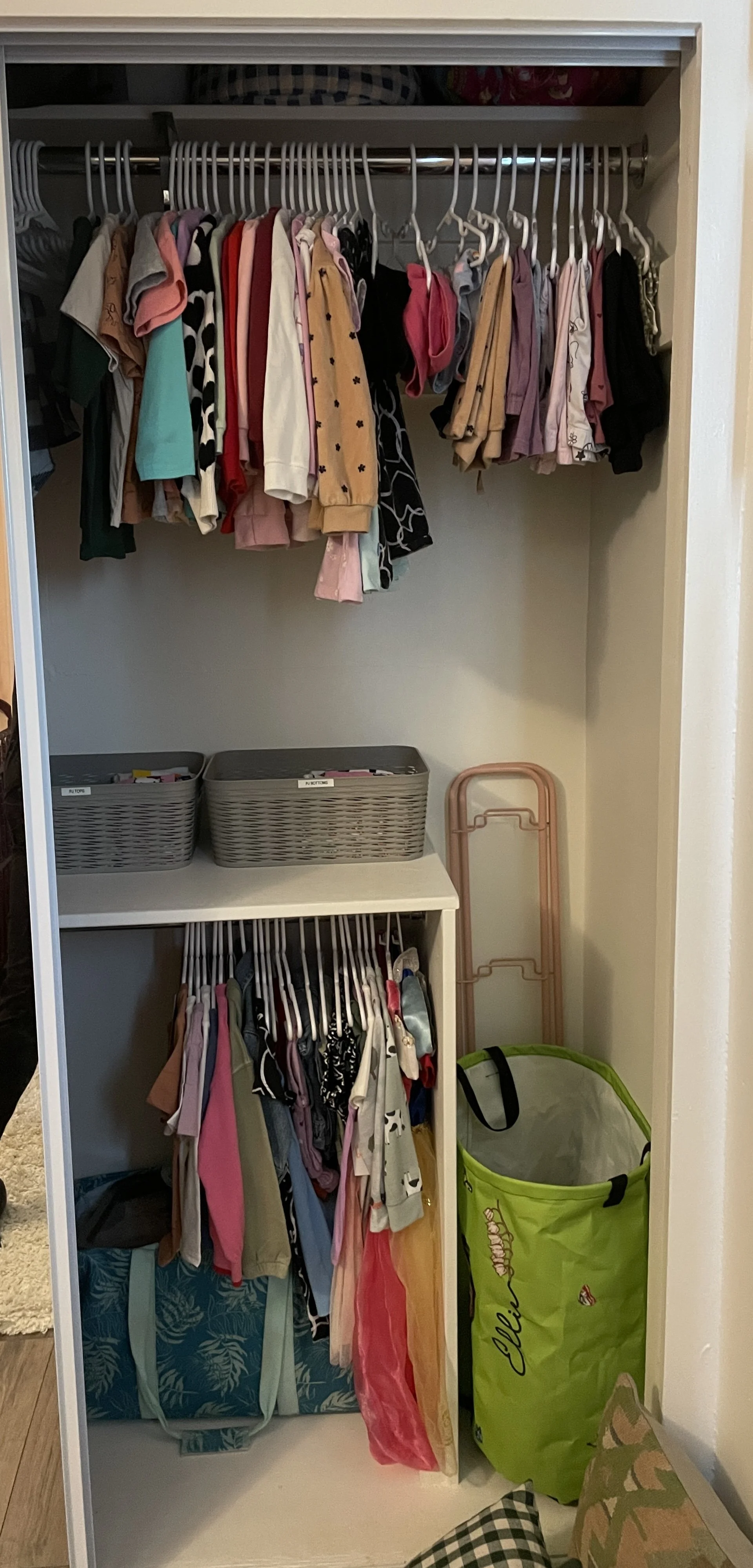 A closet with two shelves and a lower rack filled with children's clothing hung on hangers. The closet contains storage baskets on the middle shelf, a pink foldable drying rack, a green laundry tote, and a checkered cloth item in the bottom corner.