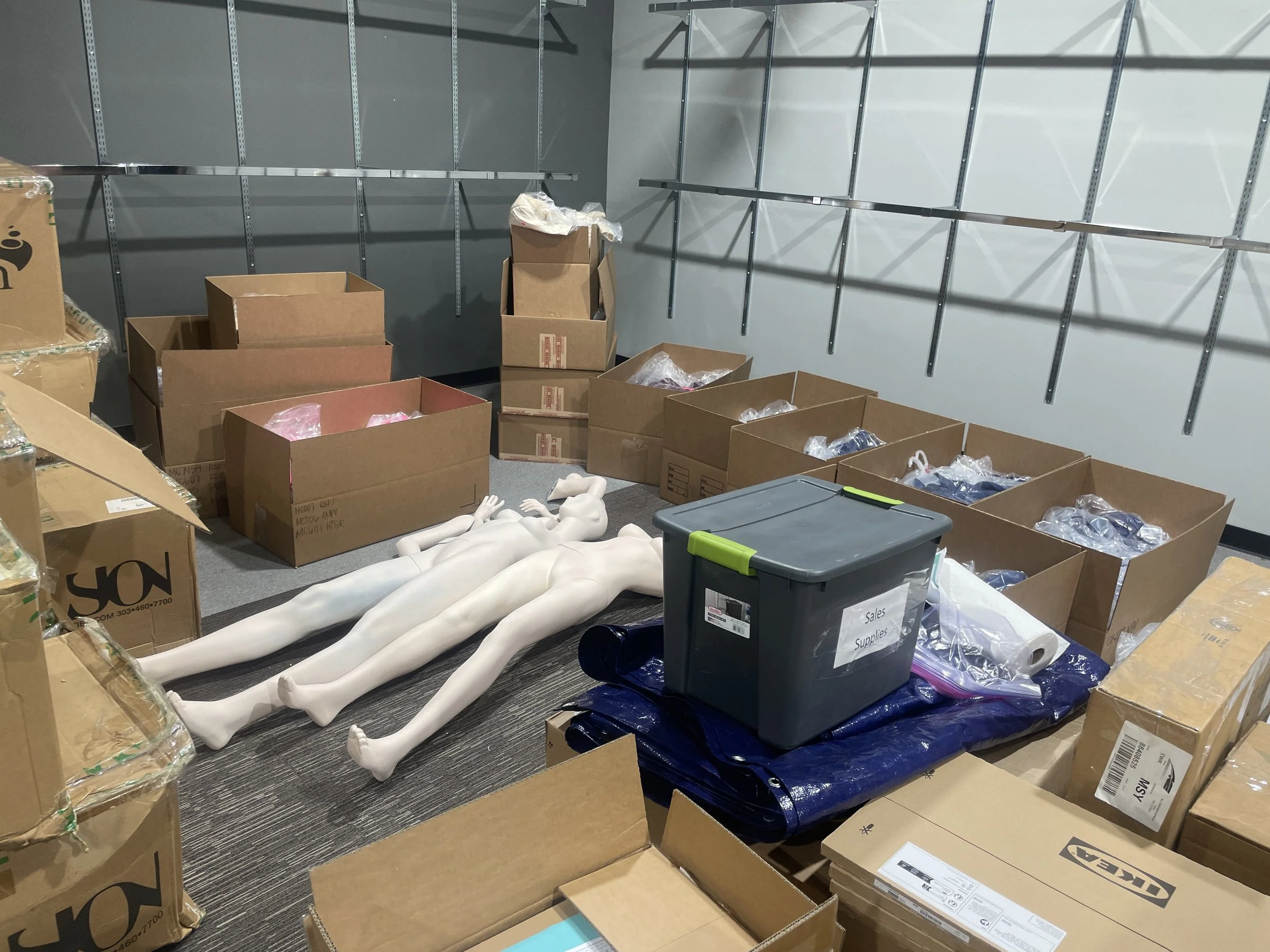 A storage room with cardboard boxes, a plastic container labeled 'Sales Supplies', a folded blue tarp, and four white mannequin limbs lying flat on the floor.