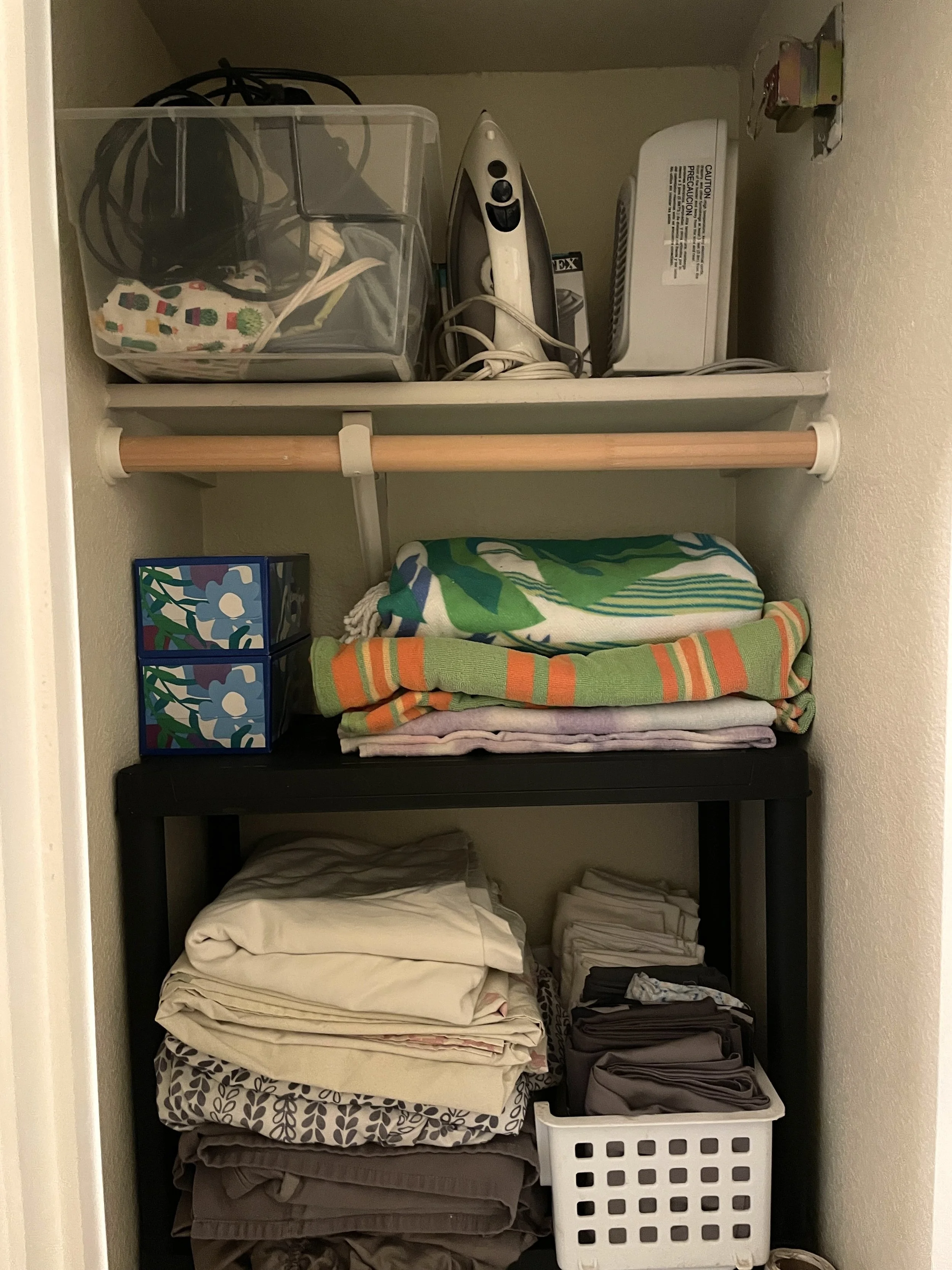 Closet with shelves containing folded clothes, a basket with clothes, an iron, and some small storage boxes.