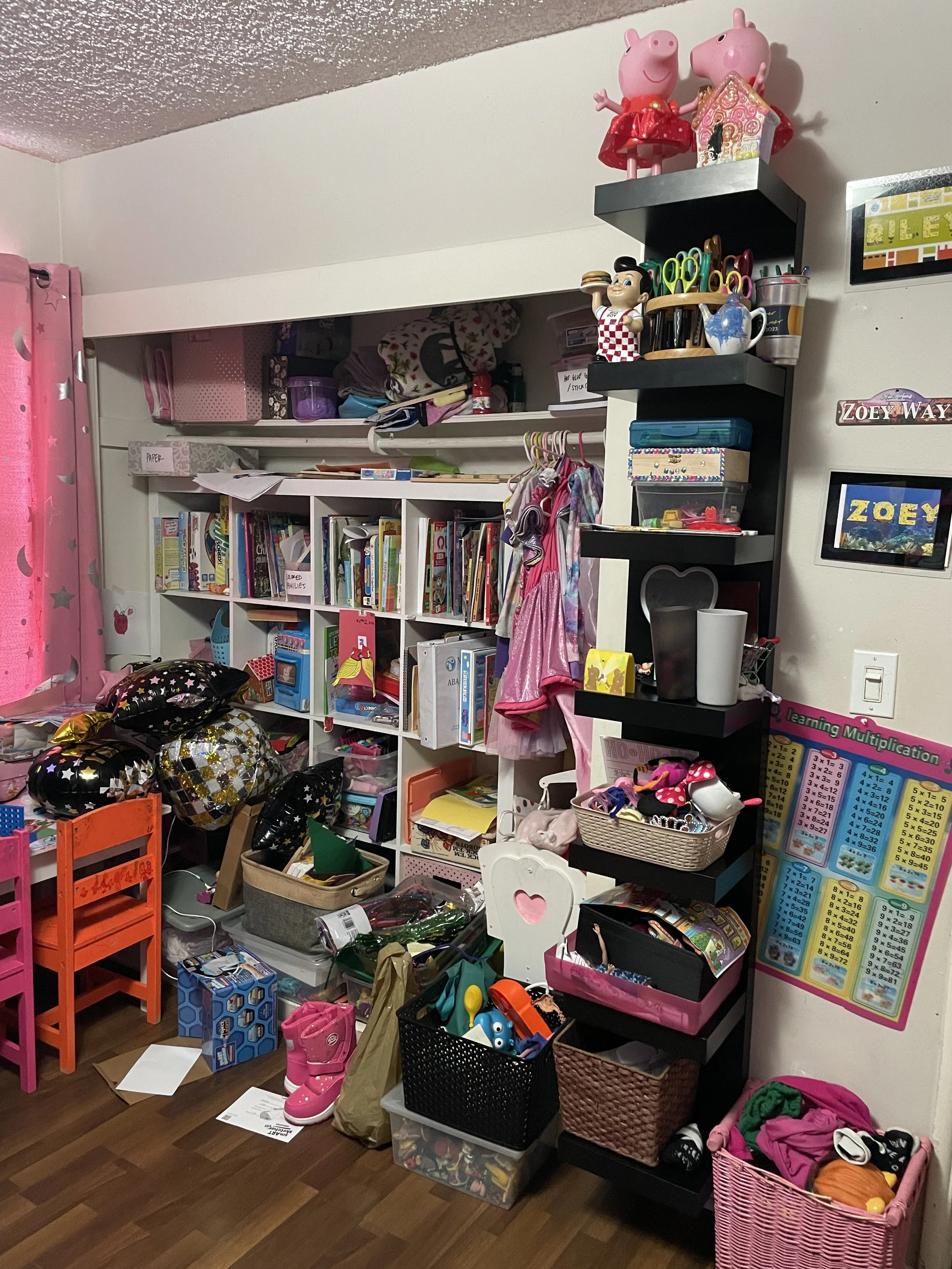 A cluttered children's room with toys, books, and clothes. There are shelves filled with toys and books, a pink curtain, and pink and orange child's chairs on the wooden floor.
