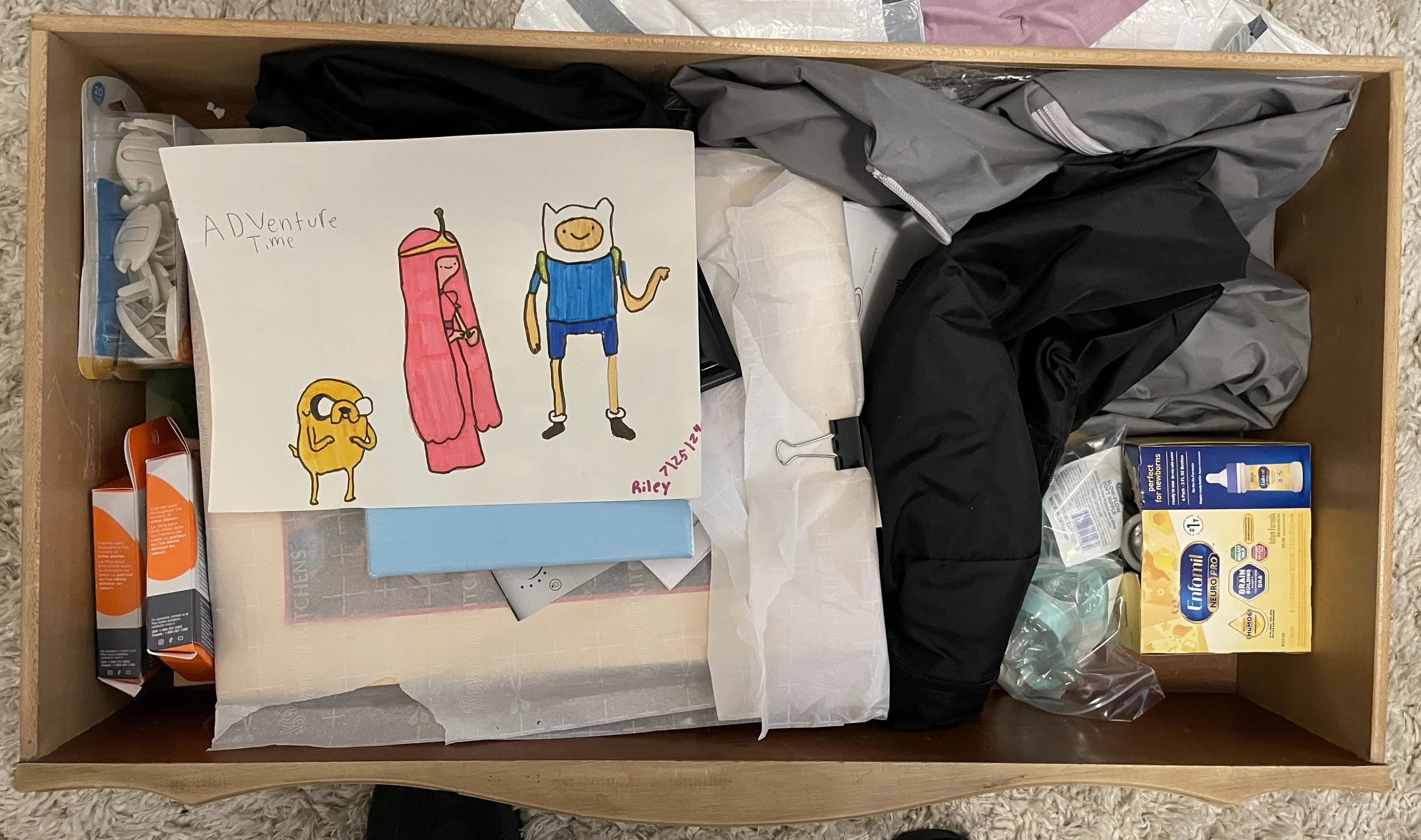 A drawer filled with various items including a hand-drawn cartoon of characters from the show Adventure Time, medication boxes, plastic bags with capsules, black and gray clothing or fabric, and a clip holding a piece of tissue paper.