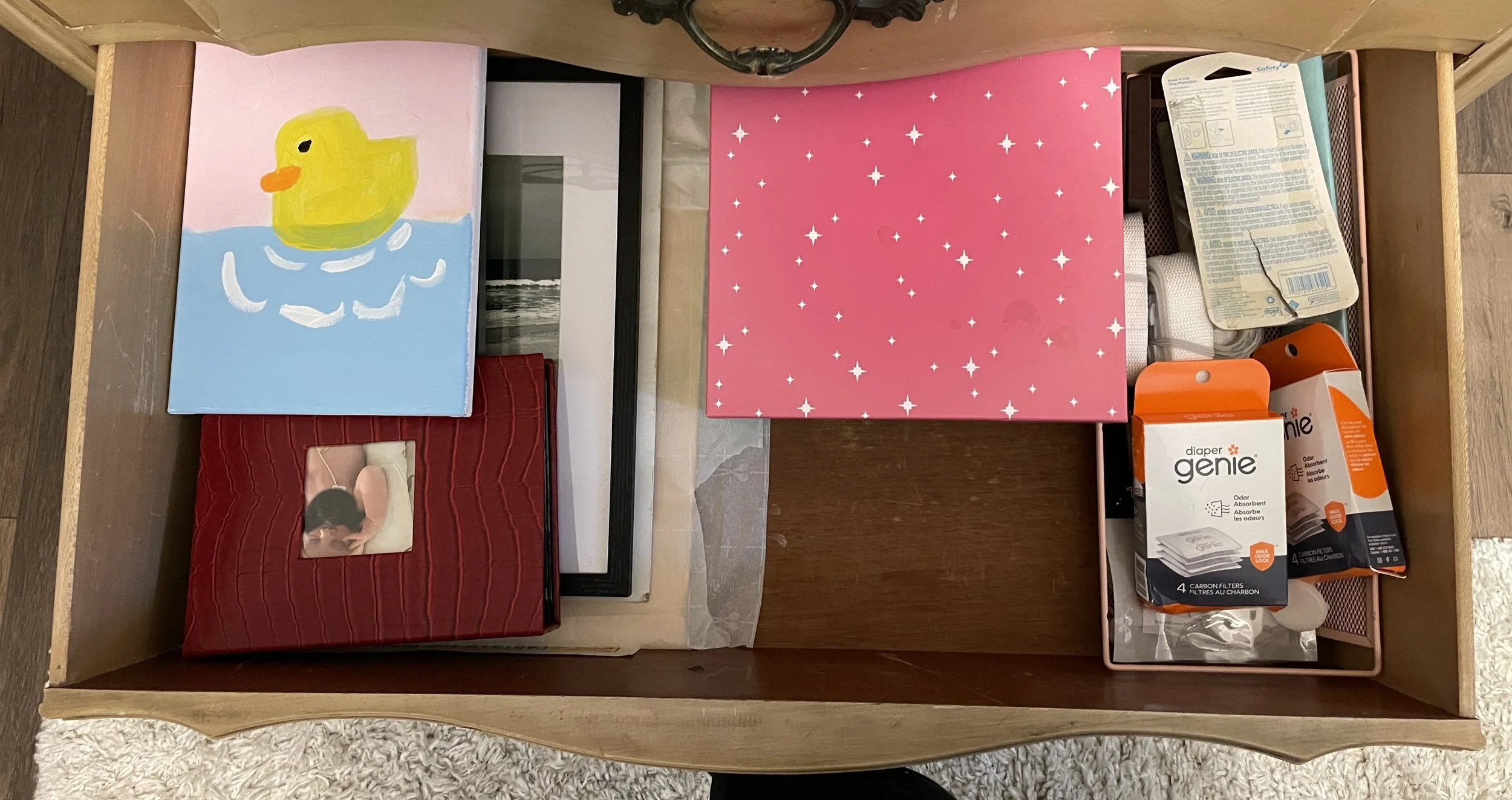 The inside of a wooden drawer containing a painting of a yellow rubber duck on blue water, a red photo album, a pink gift box with star patterns, a pack of diaper genie filters, a small container with assorted items, and a folded piece of fabric.