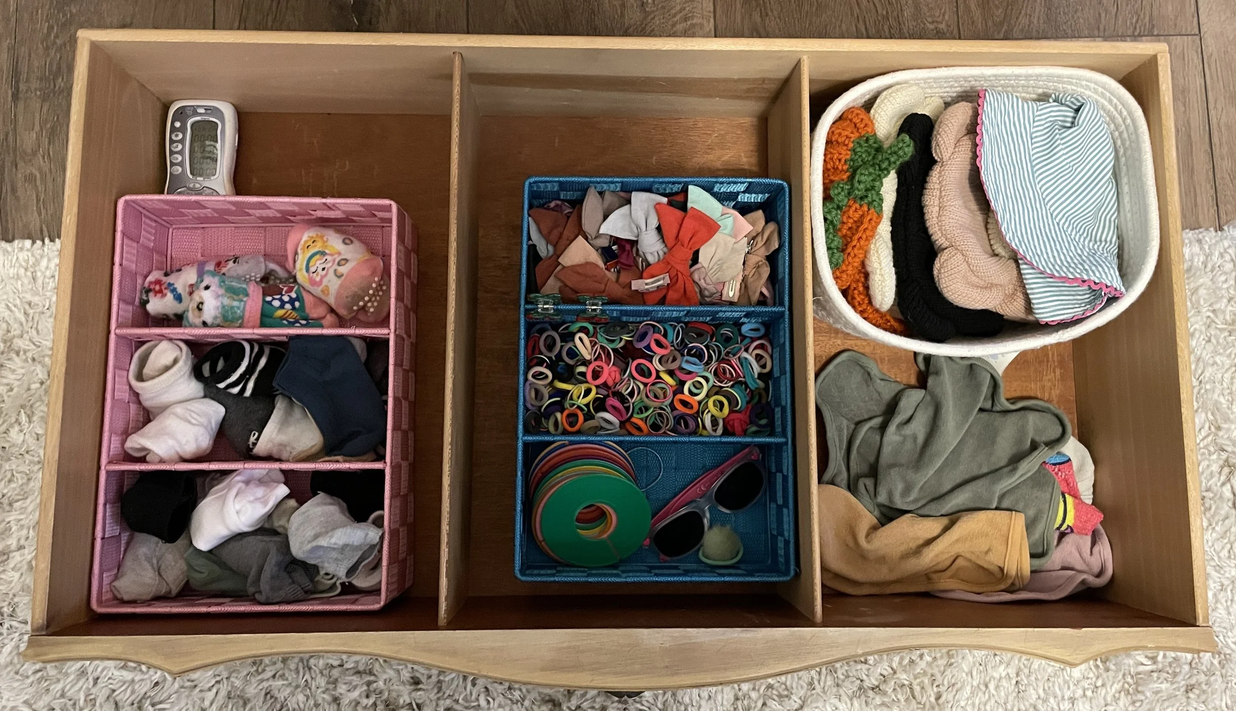 A wooden drawer divided into three sections, containing various small items. The left section holds categorized socks, the middle has colorful hair ties and small items, and the right contains folded clothes and hats.