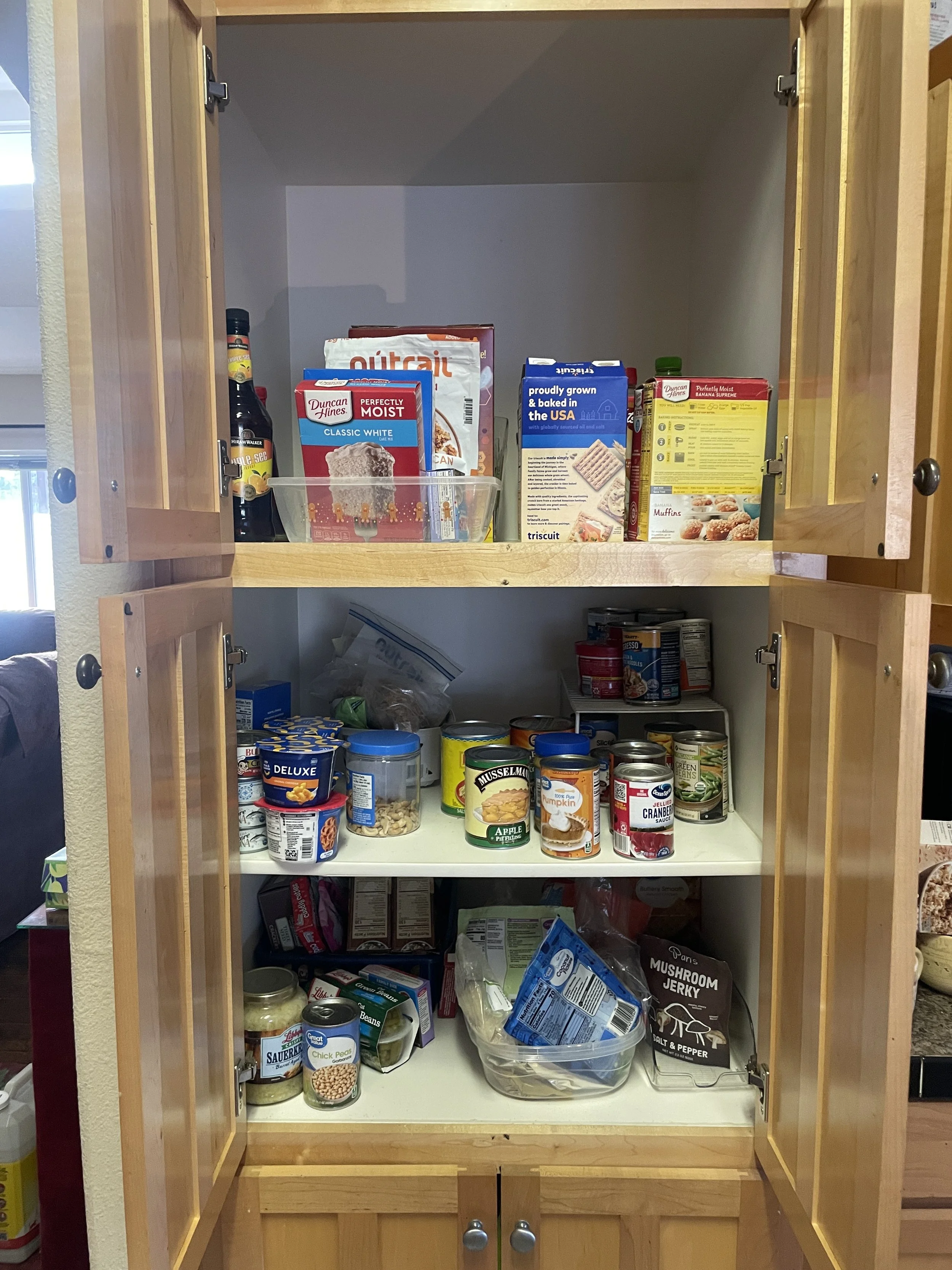 Open wooden kitchen cabinet with food items including canned goods, baking ingredients, and snacks on the shelves.
