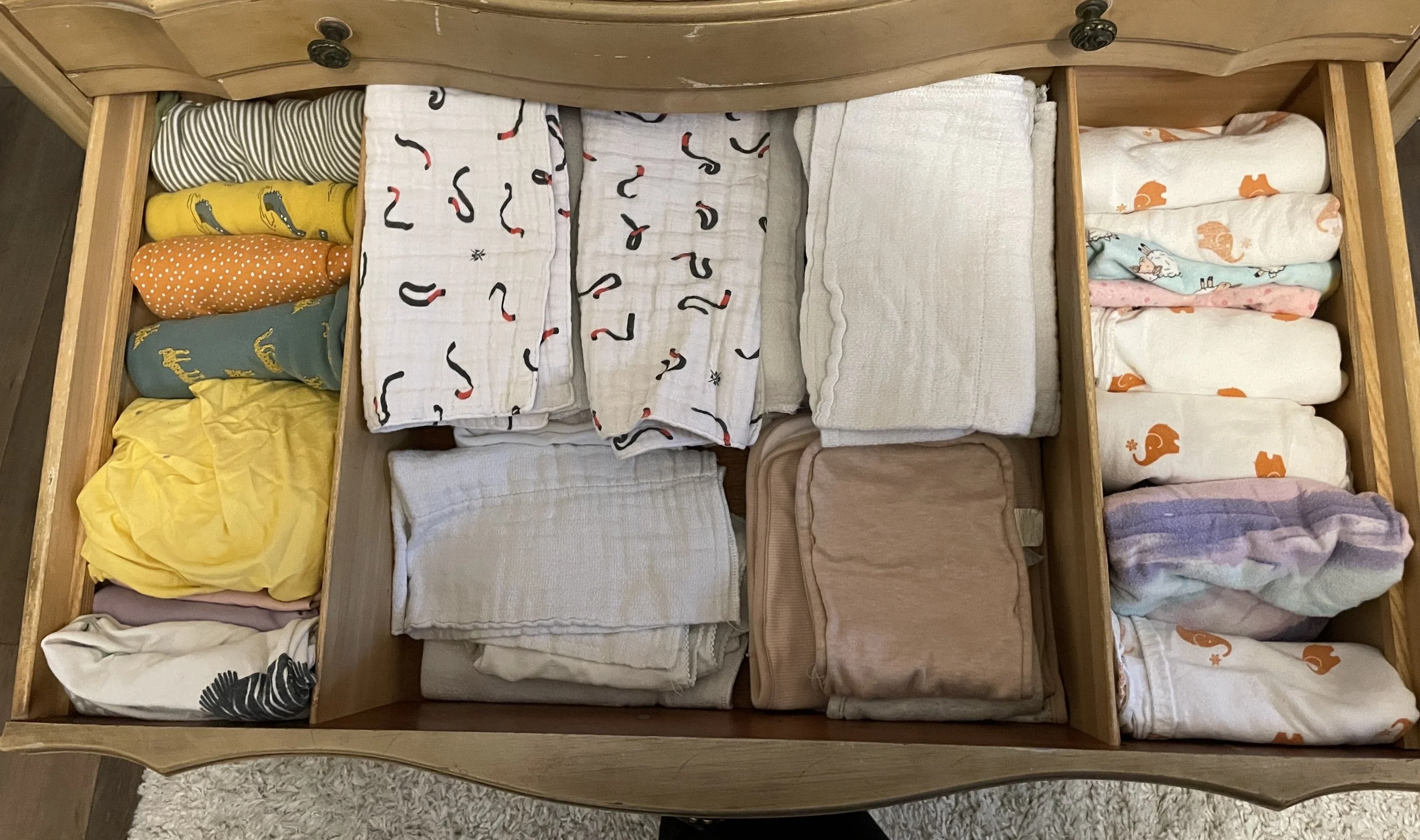 Open wooden drawer containing neatly folded baby clothes and blankets, with various colors and patterns, including those with dinosaur and whale designs.