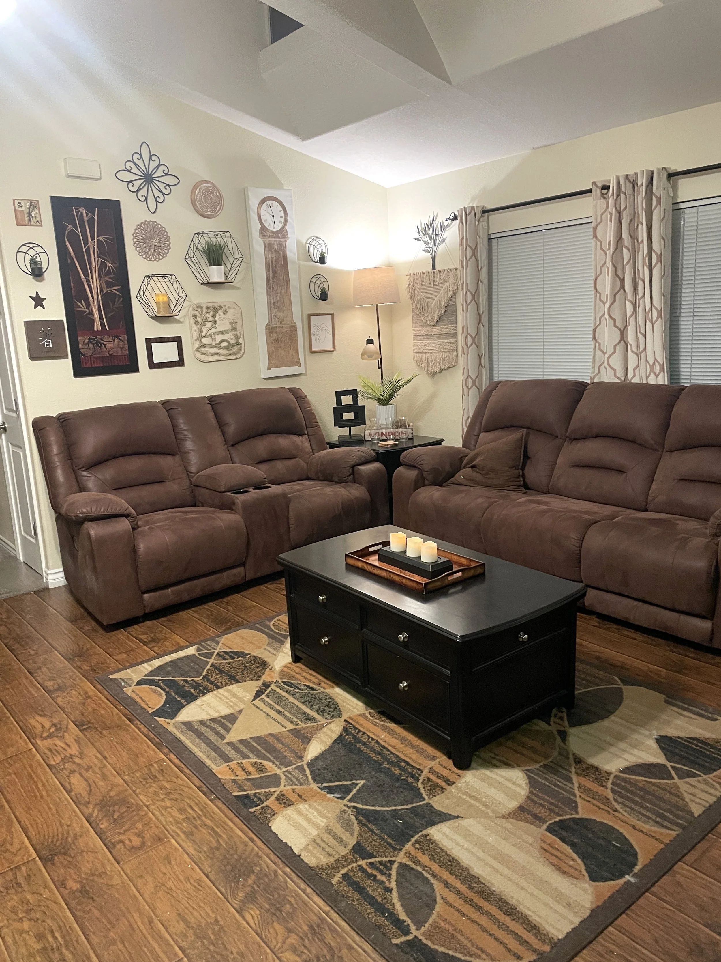 Living room with two brown leather recliners, a black coffee table with candles, a patterned area rug, a wall gallery with various decor items, a floor lamp, and double windows with patterned curtains.