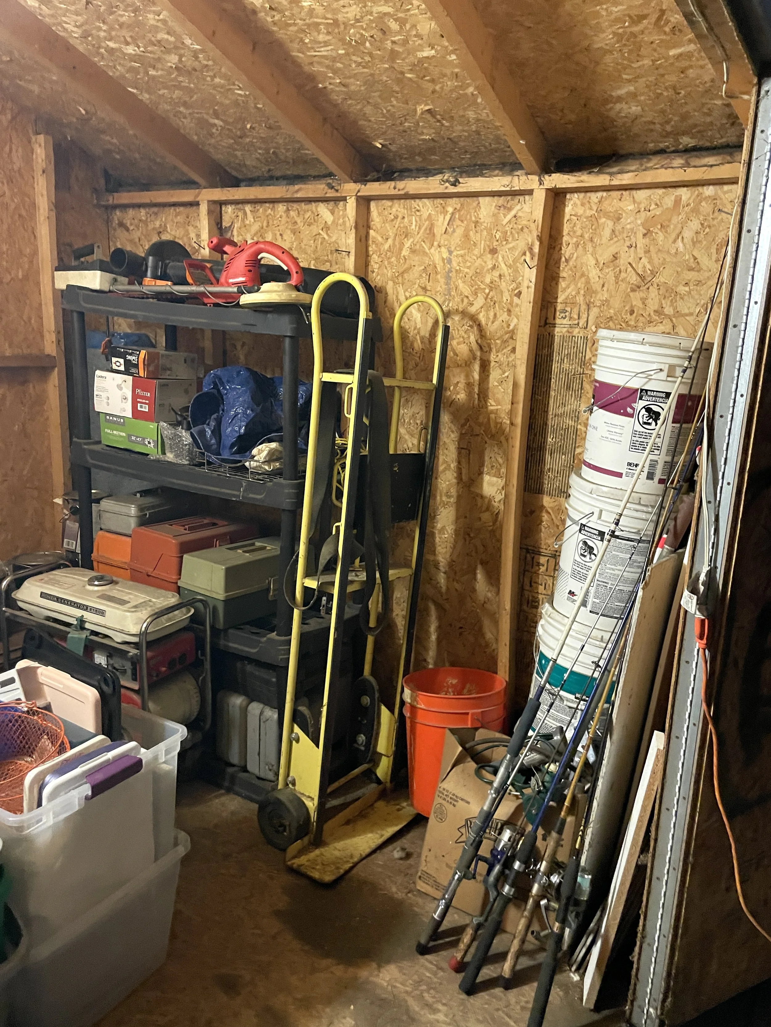 A garage wall with shelving and storage, including power tools, hand tools, and fishing rods, with plywood and OSB wall panels.