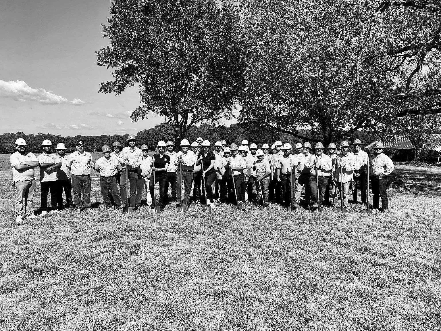 A large group of people standing outdoors in front of construction equipment, including several yellow excavators, with a young girl sitting on one of the machines. The group appears to be posing for a photo.