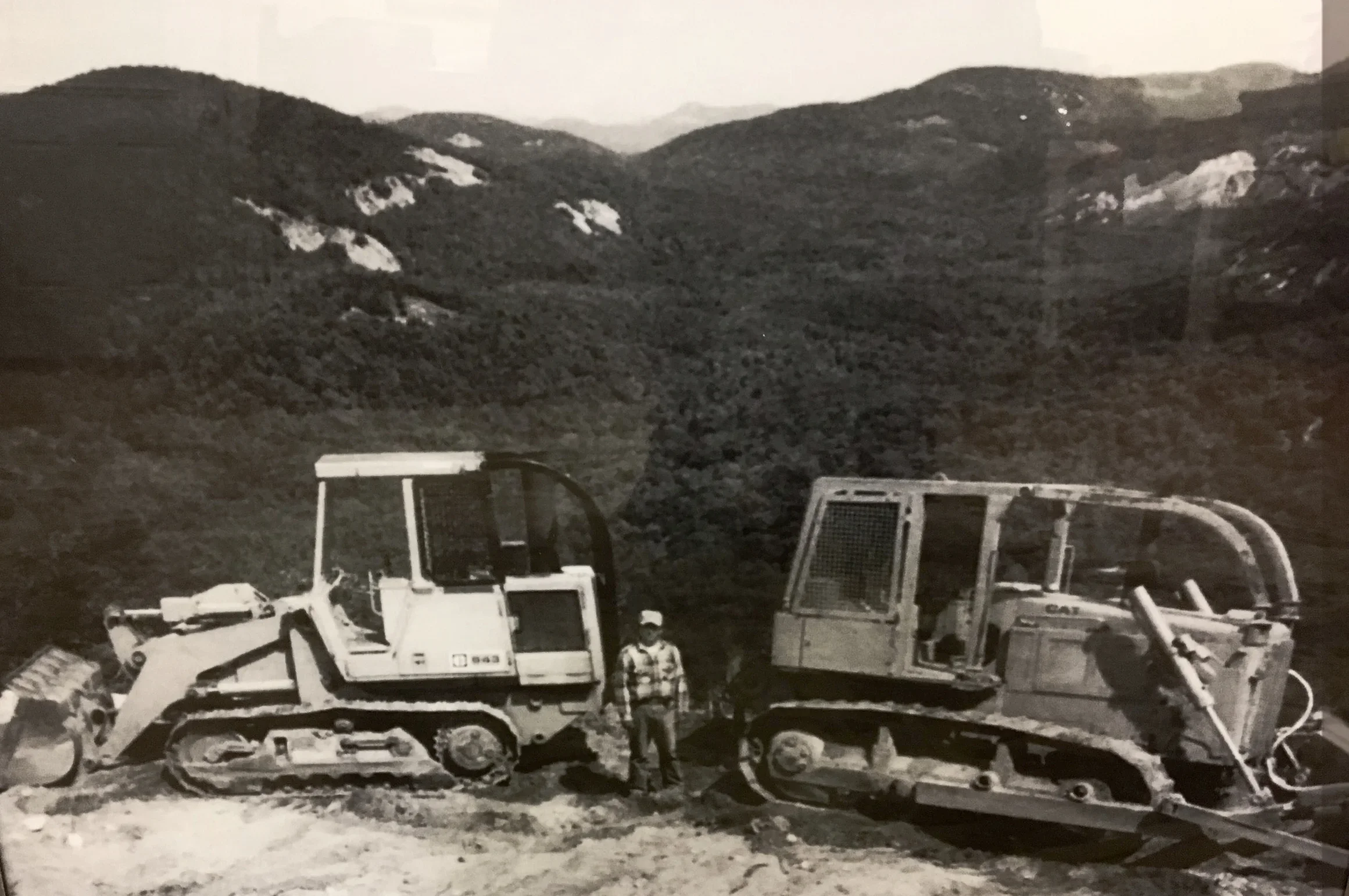 Construction site with a yellow CAT bulldozer, a worker in an orange shirt and safety helmet using a leveling tool, red dirt ground, safety barriers, and trees in the background under a clear blue sky.