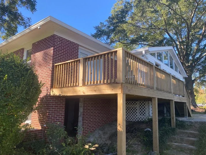 A brick house with a newly built wooden deck or balcony extending from the second floor, with a railing and a supporting post underneath, surrounded by trees and greenery.