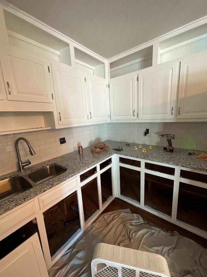 Kitchen undergoing renovation with white upper cabinets and glass-front lower cabinets, granite countertops, a double sink, and construction tools and materials on the counter.