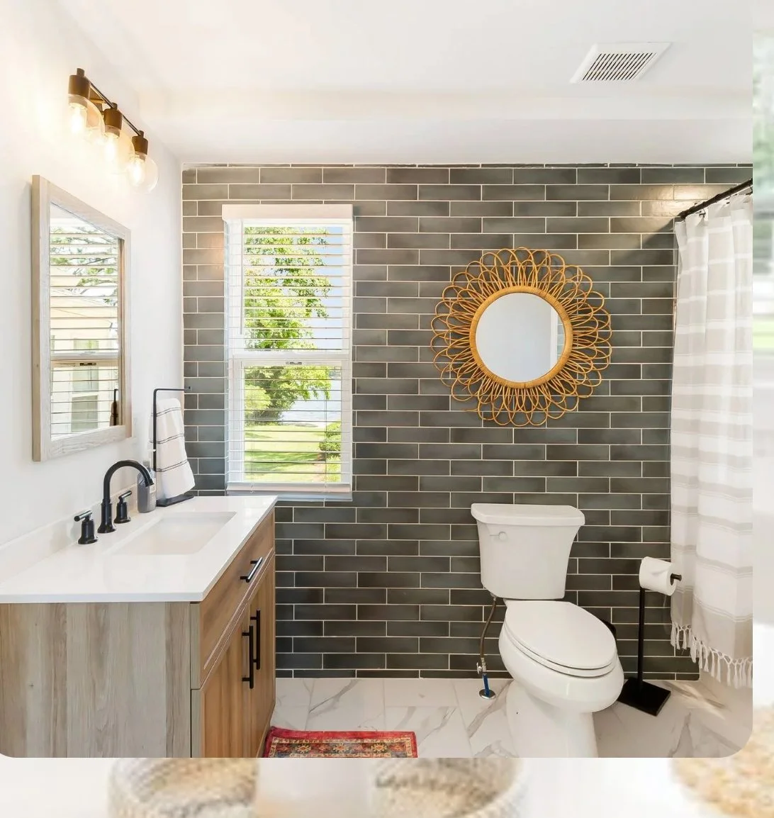 Bathroom with gray brick wall, white toilet, wooden vanity with black fixtures, round mirror with woven frame, window with blinds, and shower curtain.