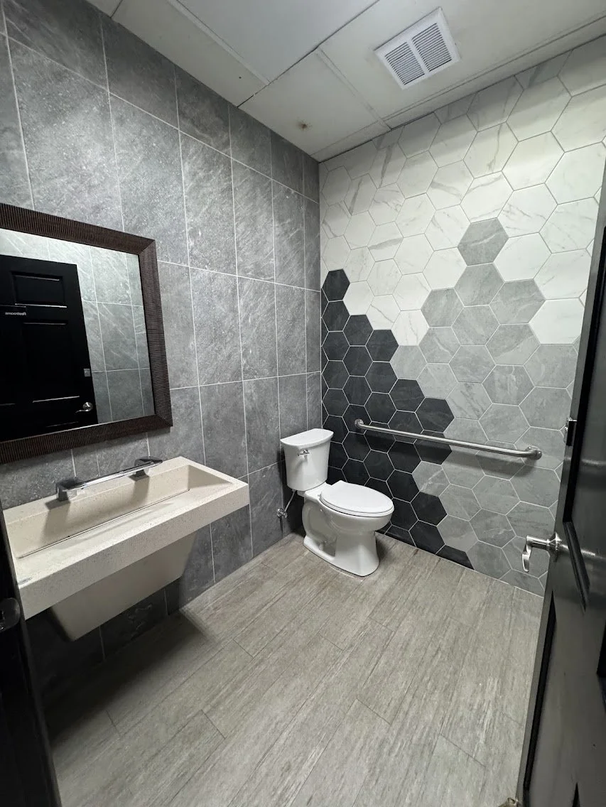 Empty public restroom with gray tiled walls, hexagon patterned accent wall, gray floor tiles, a white toilet, a rectangular beige sink, a large mirror, and a metal grab bar.