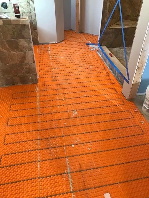 Undergoing installation of radiant floor heating with orange heat cable mats on floor, in a bathroom under construction.