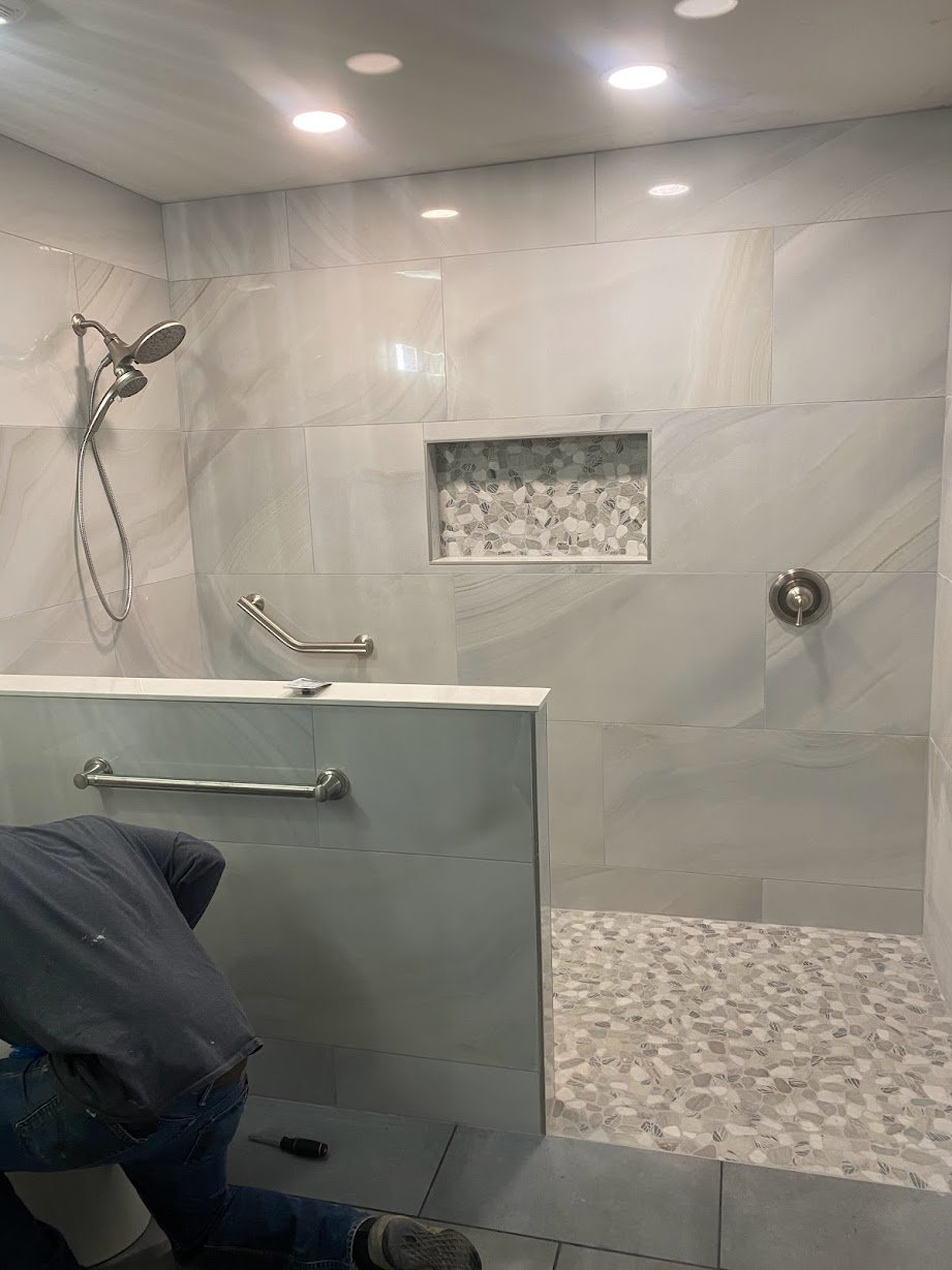 A person installing a grab bar in a walk-in shower with light-colored wall tiles, a built-in niche with mosaic tiles, and a pebble floor.