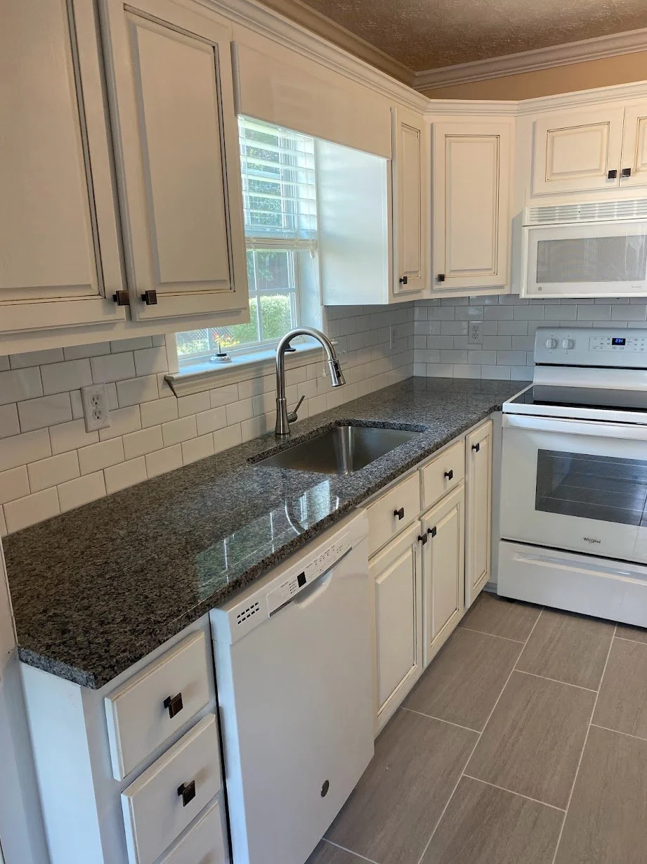 Kitchen with white cabinets, granite countertop, white subway tile backsplash, window above sink, stainless steel faucet, white dishwasher, and white oven and microwave.