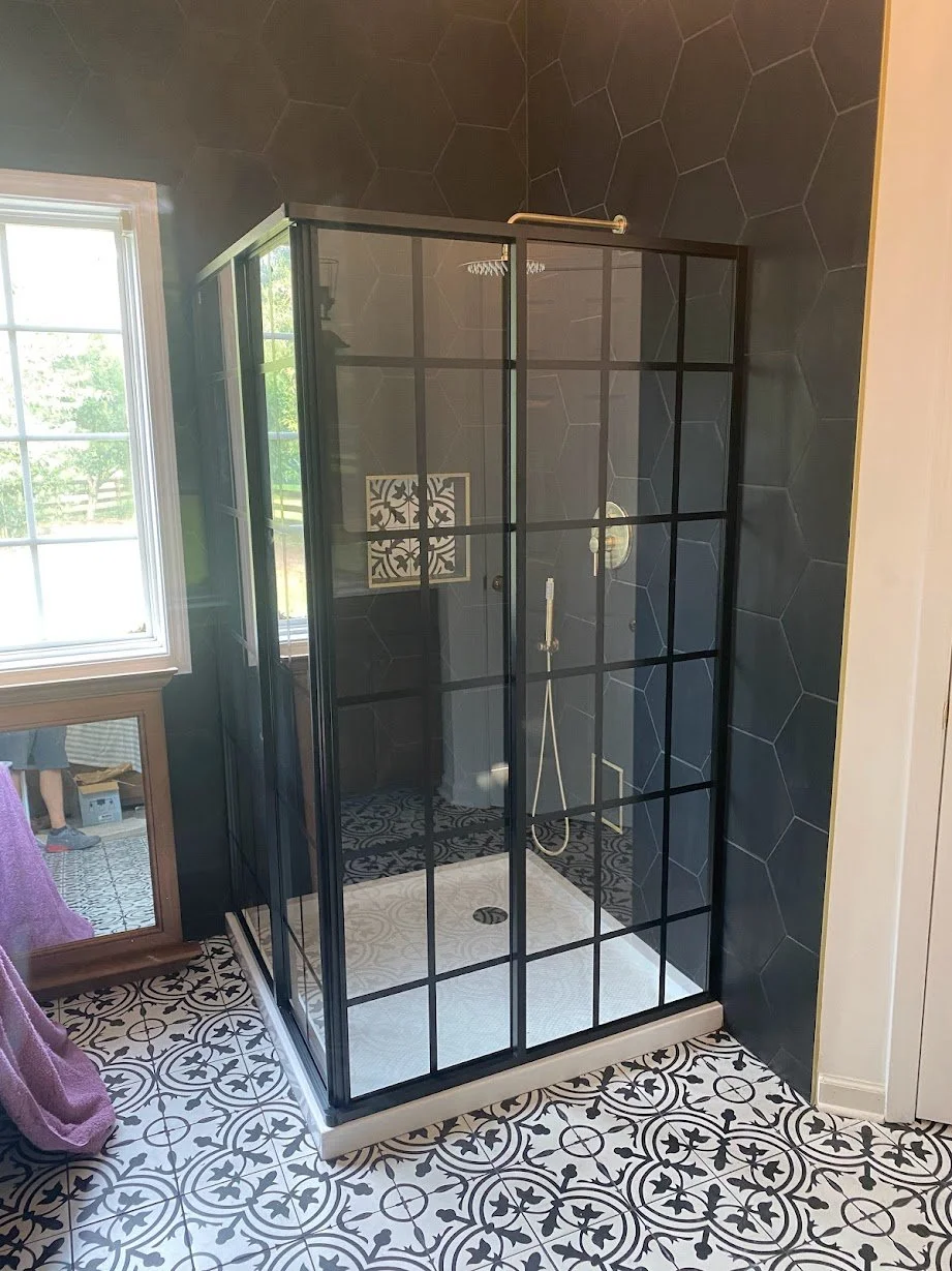 A modern shower with black grid-framed glass panels, dark hexagonal tiled walls, patterned black and white floor tiles, and a window reflecting greenery outside.