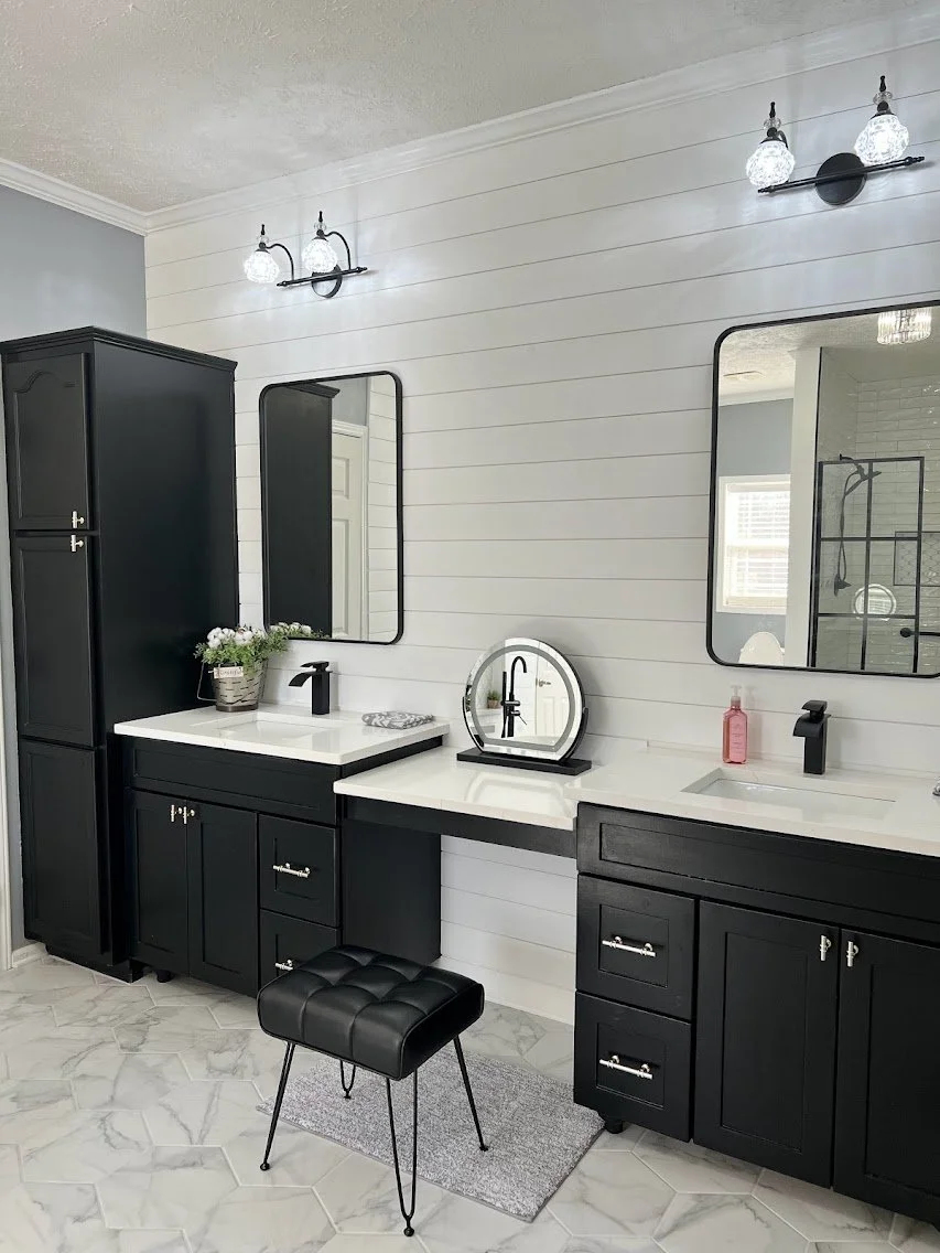 A modern bathroom vanity area with two black and white sinks, two rectangular mirrors, black cabinetry, a black storage cabinet, a black padded stool, and wall-mounted light fixtures with three bulbs each. There are decorative items including a small
