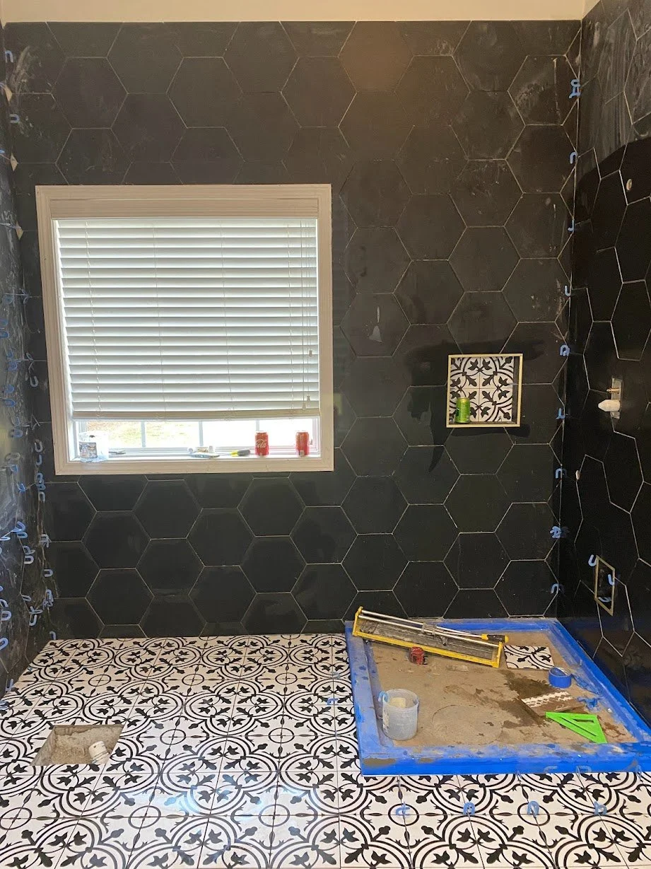 Bathroom under renovation with black hexagonal tile walls, patterned floor tiles, a window with blinds, and construction tools and materials on the floor, including a blue framed shower base.