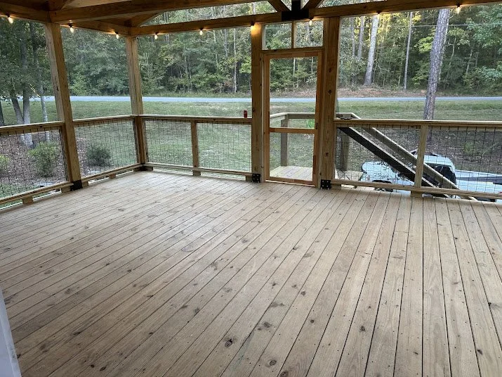 Empty wooden deck with railing, surrounded by trees and overlooking a road and parked vehicle.