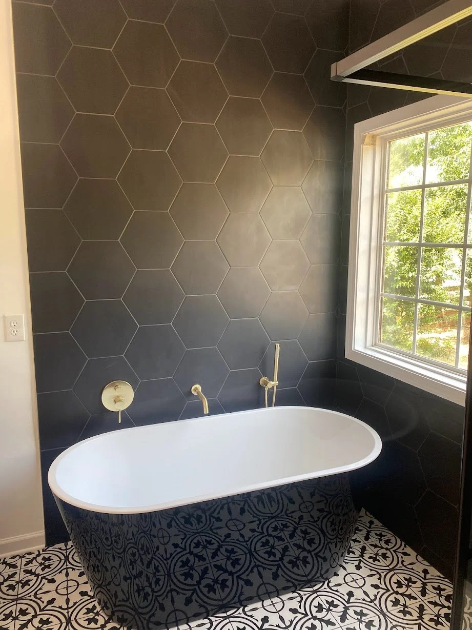 Bathroom with a bathtub, dark hexagonal wall tiles, patterned floor tiles, and a window with greenery outside.