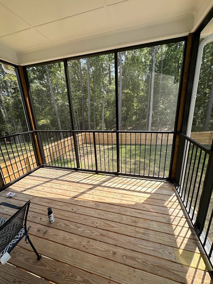 A screened porch with wooden floor, black railing, and a view of a backyard with grass and trees.