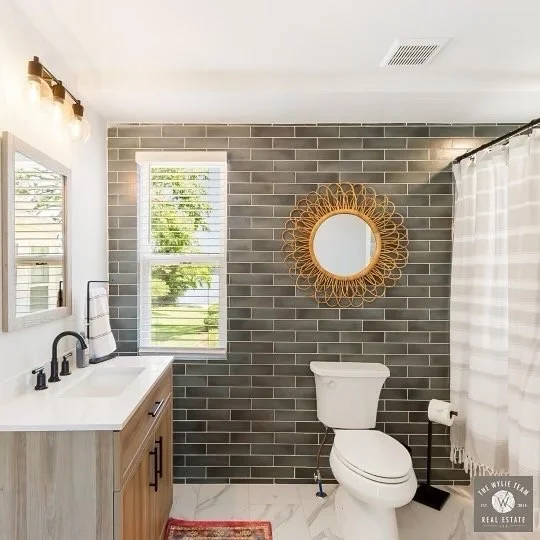 Modern bathroom with gray subway tiles on the wall, a white toilet, a window with blinds, a vanity with a black faucet, a mirror, a decorative round mirror, and a checkered shower curtain.
