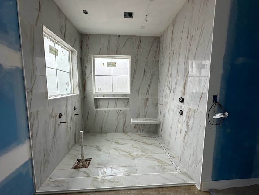 A bathroom under construction with walls and floor covered in white marble tiles with gray and gold veining. There are two large windows on the back wall, a small built-in shelf under one window, and a hole in the floor for plumbing. Exposed wiring a