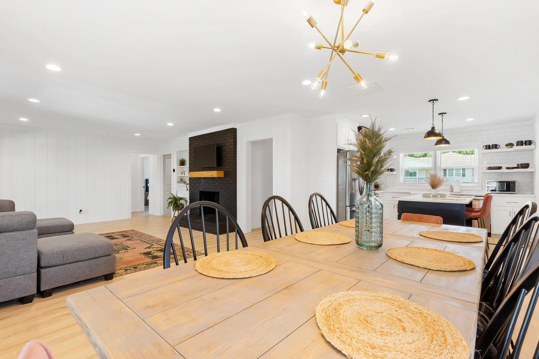 Open concept living and dining area with a wooden dining table, woven placemats, a glass vase with greenery, a gray sofa, a black brick fireplace with a mounted TV, kitchen with white cabinets, black accents, and open shelving, hardwood floors, large windows, and modern lighting fixtures.