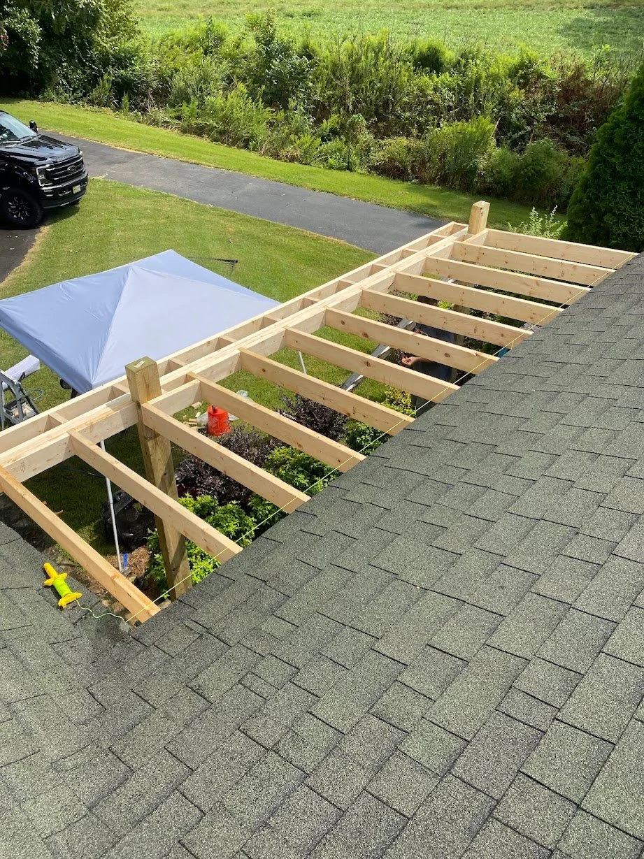 A house with its roof partially stripped to expose the wooden framing underneath, while a black truck is parked on a driveway nearby. A white canopy tent is set up behind the house, and tools are visible on the roof and in the yard.