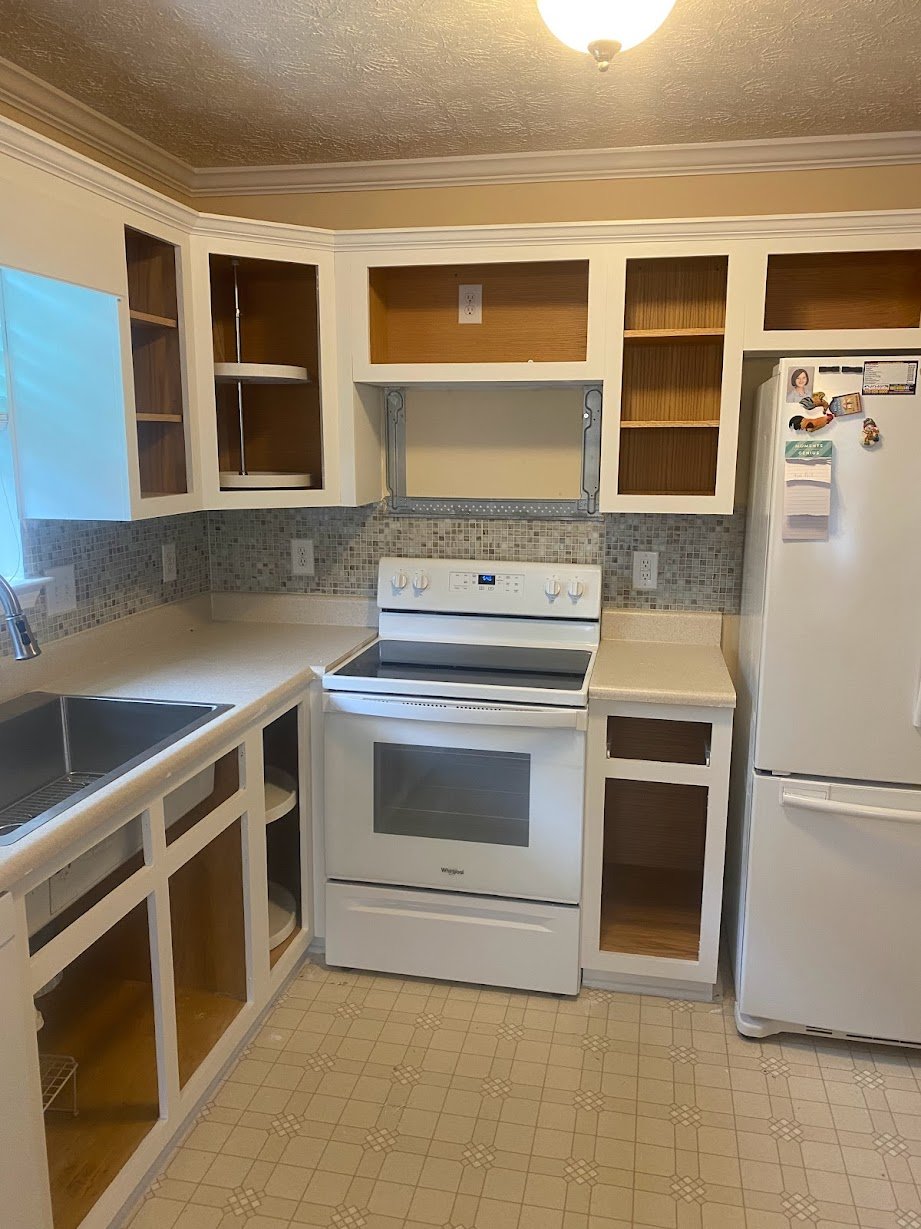 Empty kitchen with white cabinets, a white oven, a refrigerator with magnets, beige countertops, a mosaic tile backsplash, a double sink, and open cabinet shelves
