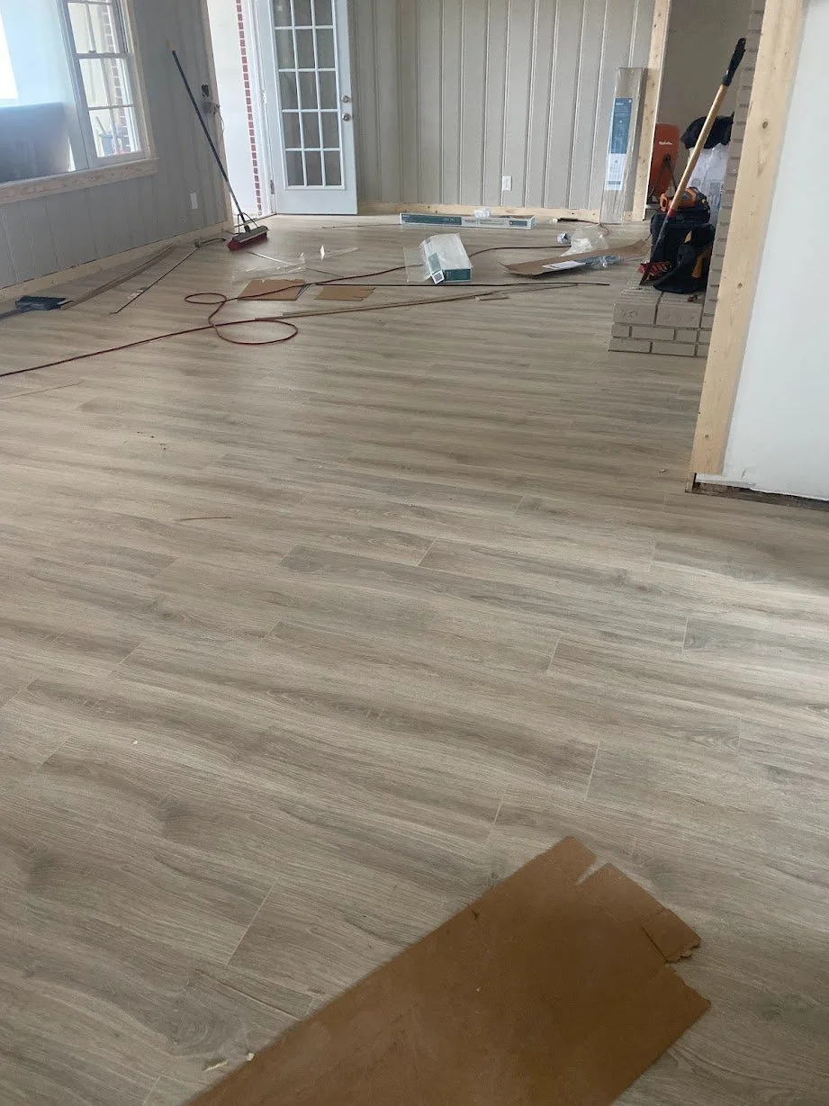 An unfinished room with newly installed light wood flooring, construction tools, and materials scattered on the floor. Construction supplies and equipment are visible, and the room has a door with glass panes leading outside.