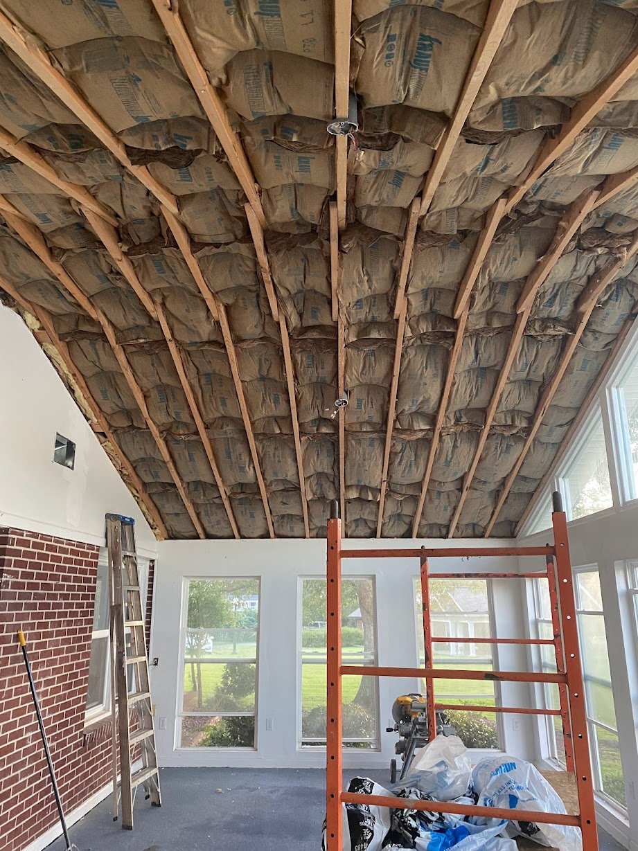 A room undergoing renovation with insulation installed in the ceiling, a ladder leaning against the wall, an orange scaffold, and construction tools and materials on the floor.