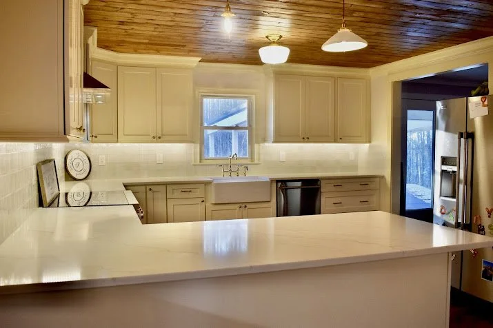 Bright kitchen with cream-colored cabinets, a white countertop, a window above the sink, and a wooden ceiling. The refrigerator and dishwasher are visible.