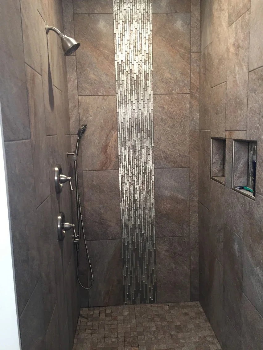 A walk-in shower with brown tile walls, featuring a vertical strip of small gray and white mosaic tiles, and a built-in niche for toiletries.