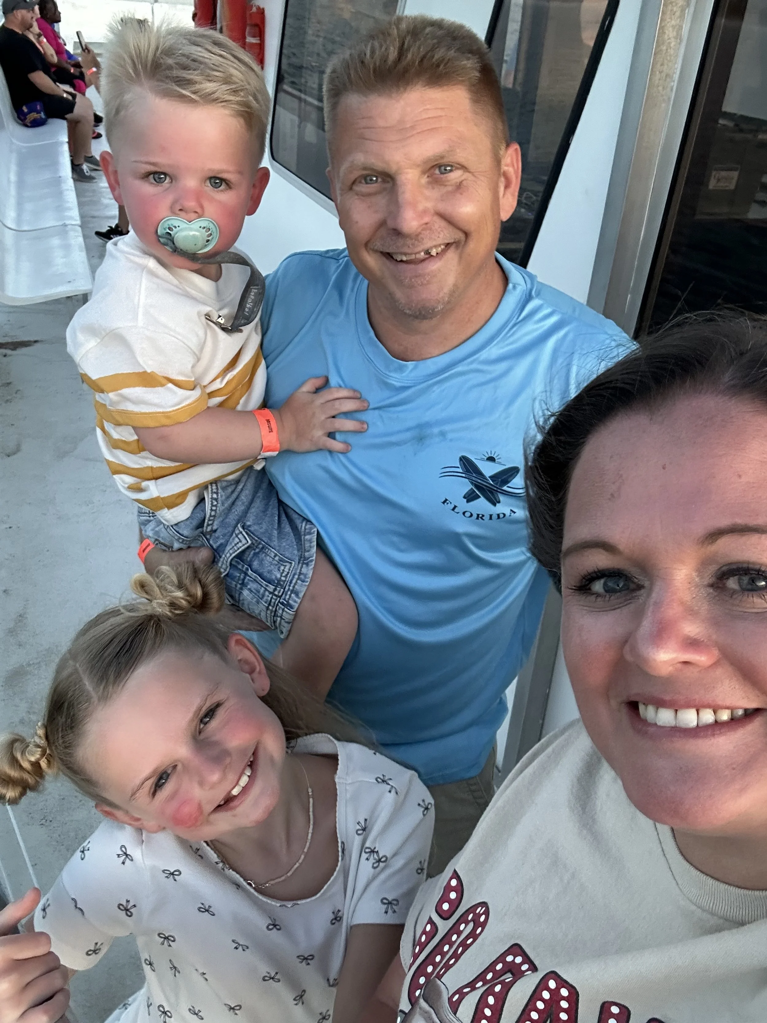 A family taking a selfie on a boat with a man, a woman, two young girls, and a young boy with a pacifier, all smiling.